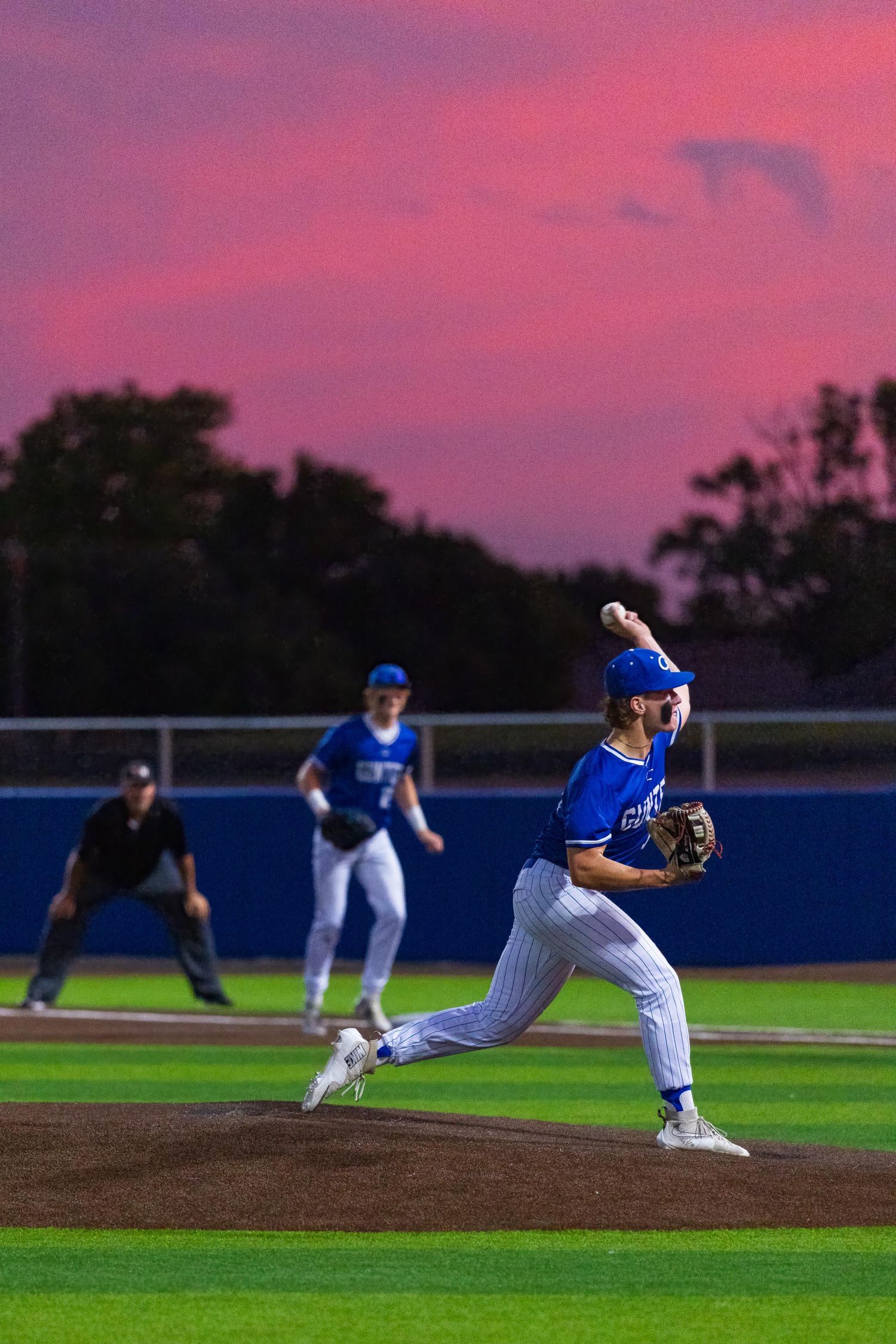 Baseball pitcher in blue uniform mid-throw at dusk with vibrant pink sunset sky backdrop on a lit field.