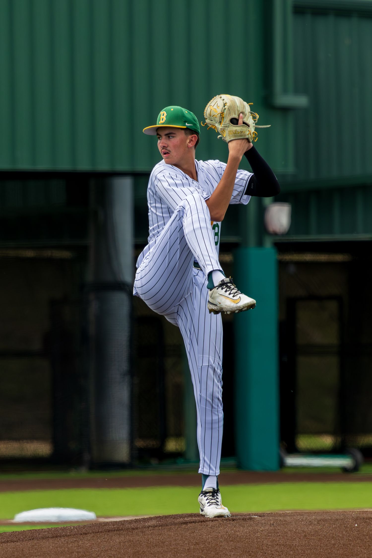 Young baseball pitcher in white pinstripe uniform and green cap winds up for a pitch on the mound.