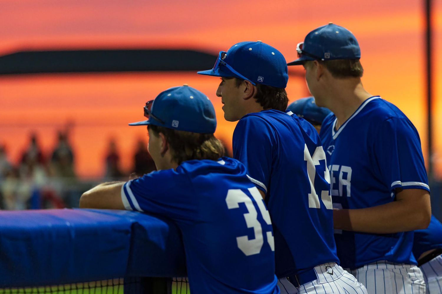 Three baseball players in blue uniforms watch from the dugout against a stunning orange and red sunset sky.