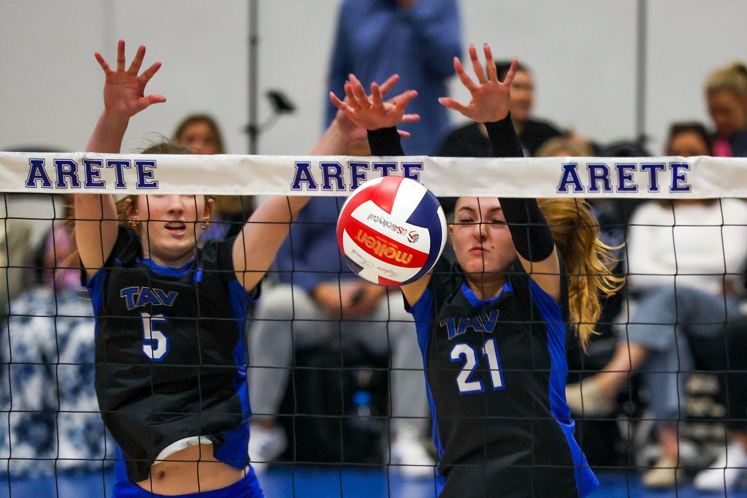 Two volleyball players in blue uniforms jump to block the ball at the net during an Arete tournament match.