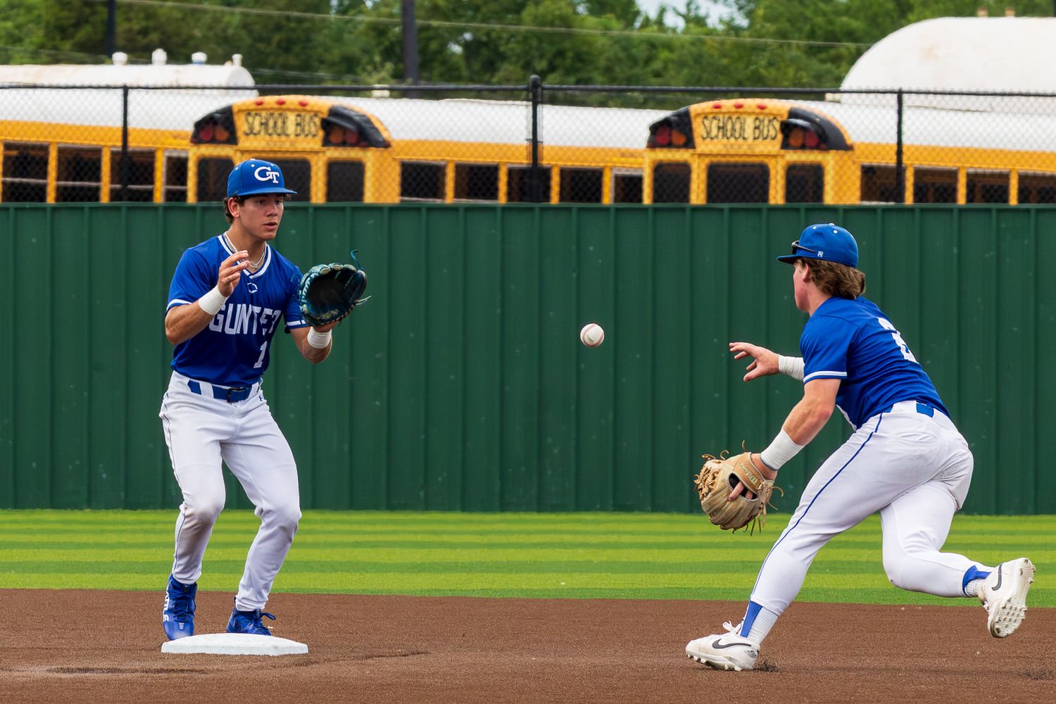Two baseball players in blue uniforms field a ball during a game, with yellow school buses visible in the background.