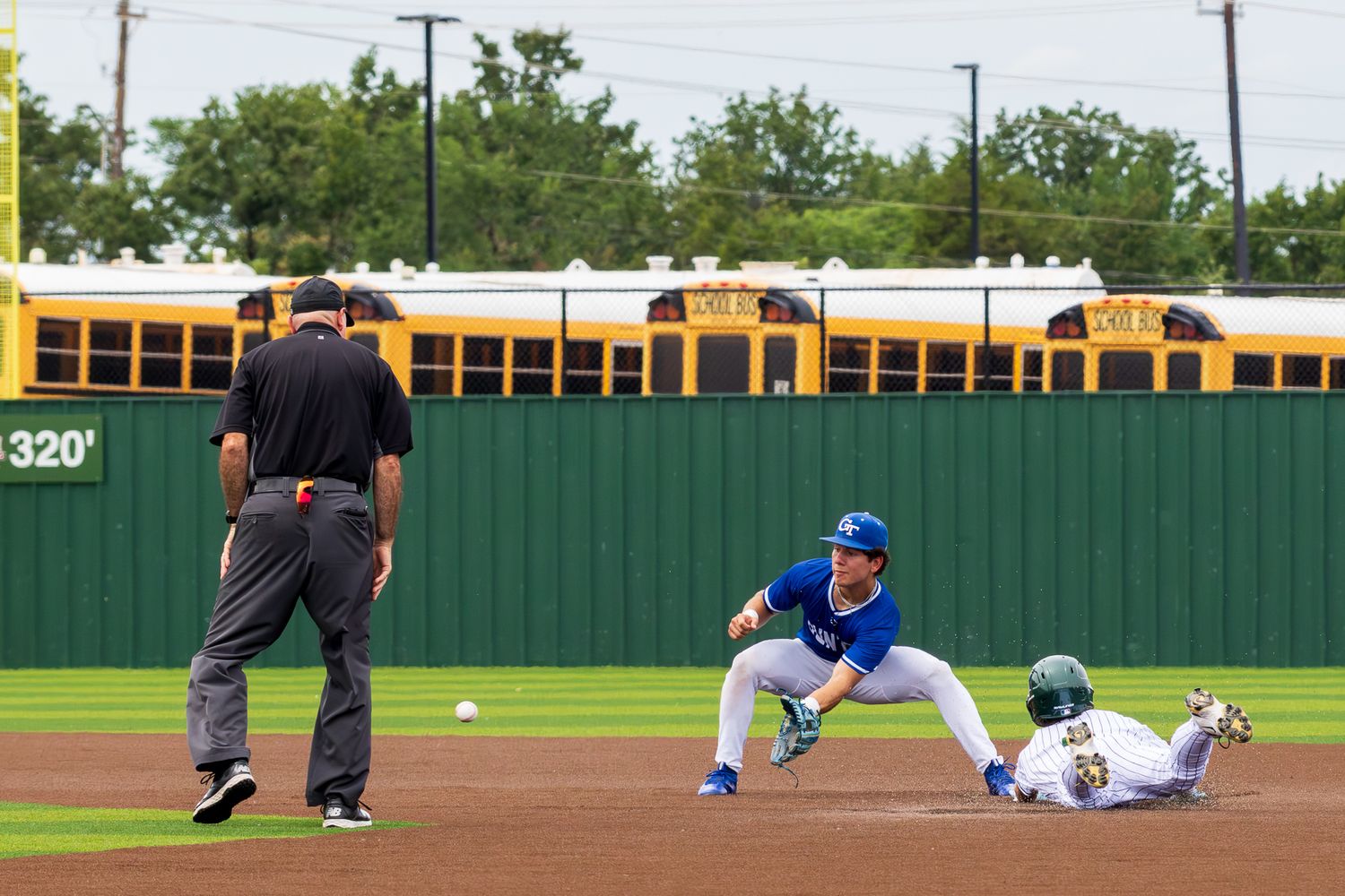 Baseball player sliding into second base as fielder catches ball, with umpire watching and school buses in background.