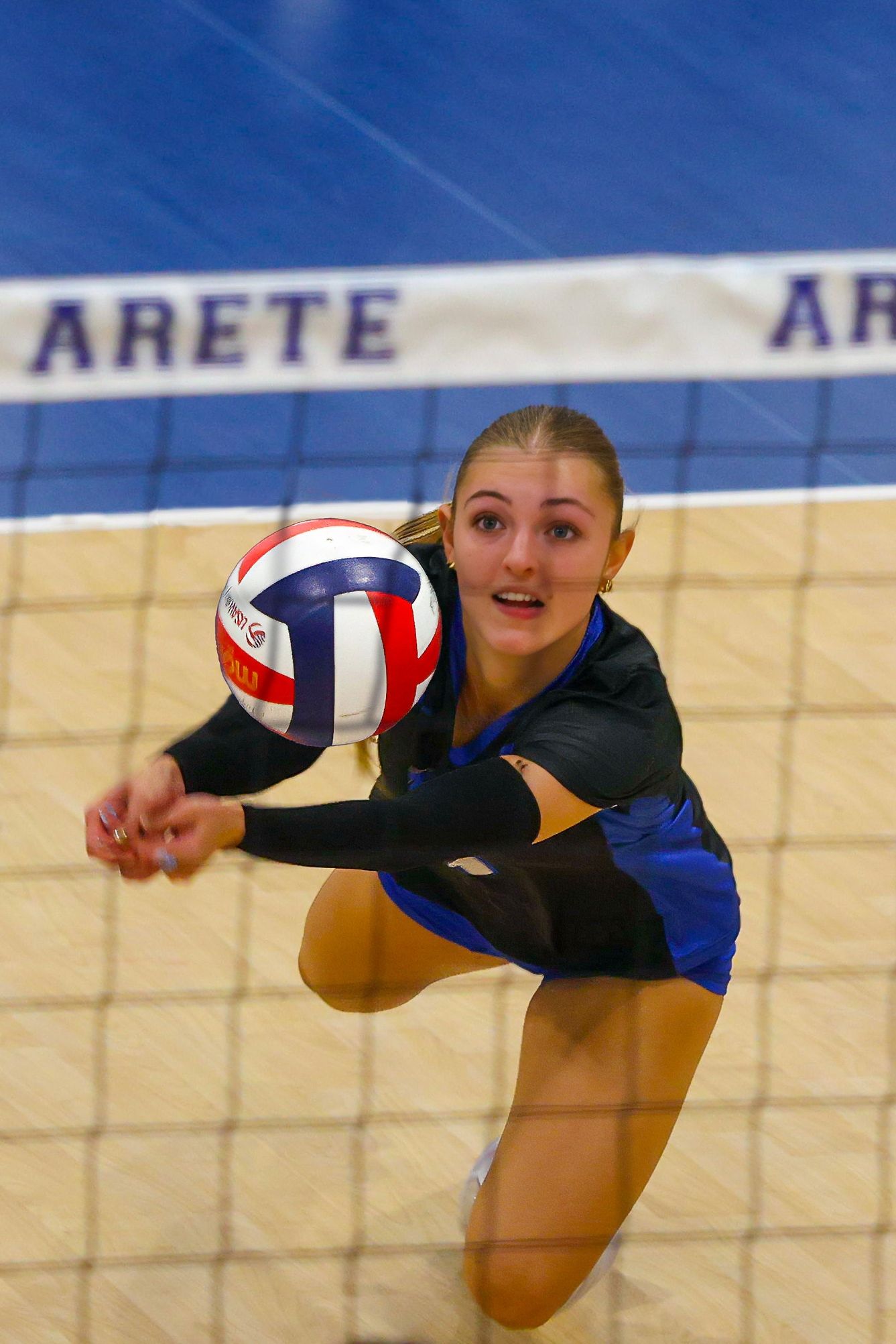 Female volleyball player performs a dig pass near the net during an indoor Arete volleyball match.