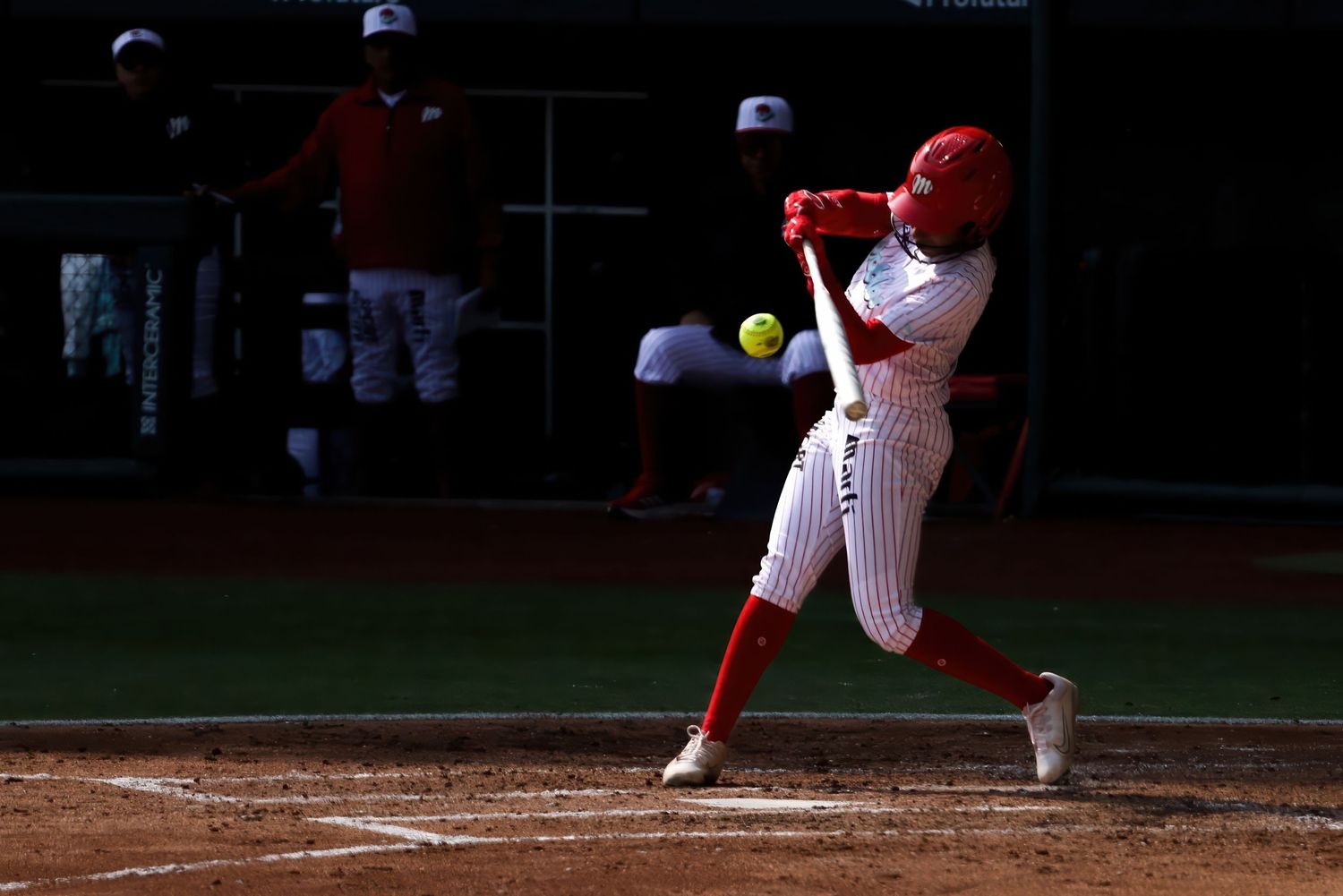 Softball batter in red uniform makes contact with yellow ball during a competitive game.