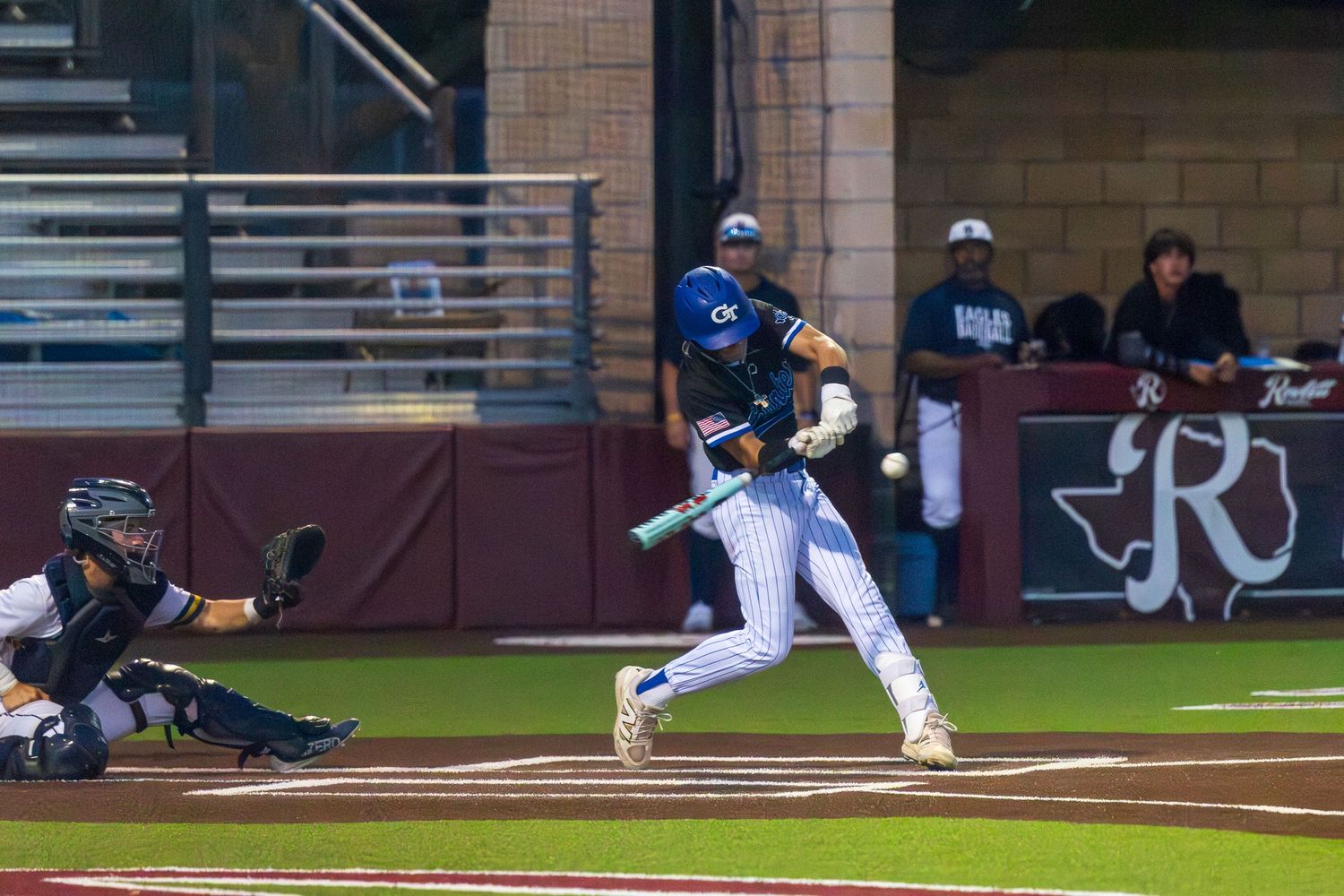 Baseball batter swings at pitch during night game while catcher crouches behind home plate at a college baseball stadium.