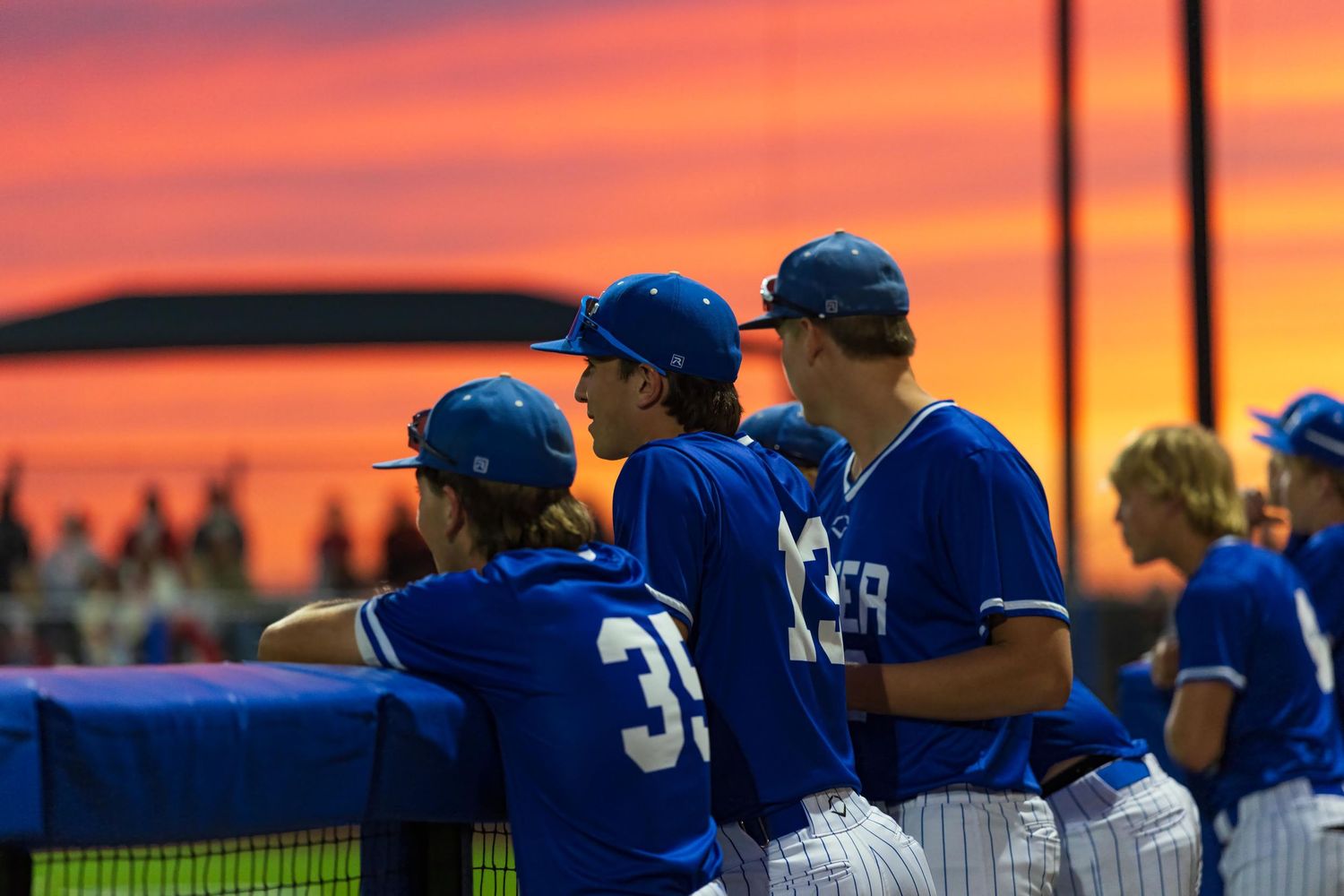 Baseball players in blue uniforms watch from the dugout against a vibrant red and orange sunset sky.