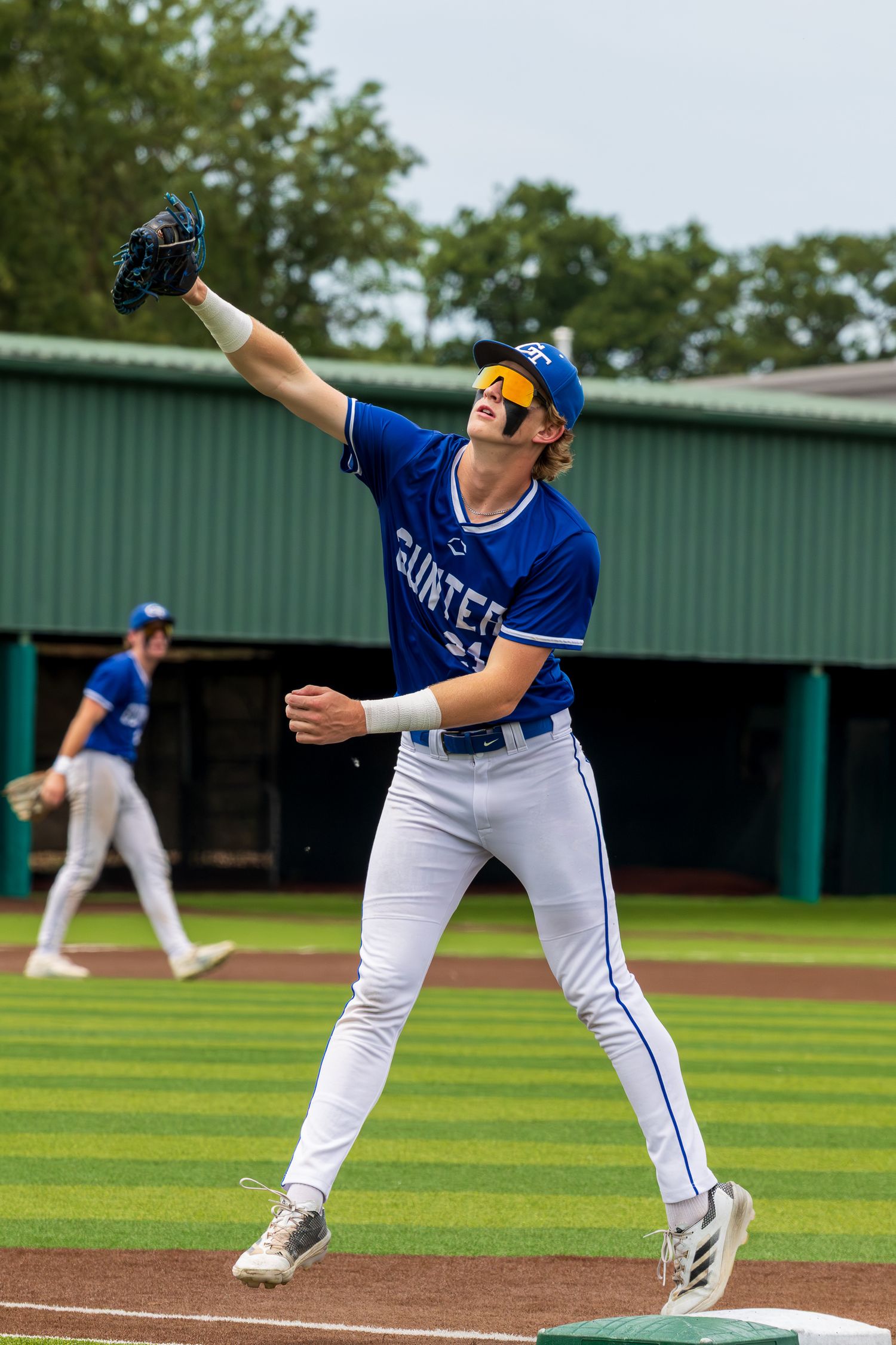 Baseball player in blue Gunter jersey reaches up to catch a fly ball at first base during a game.