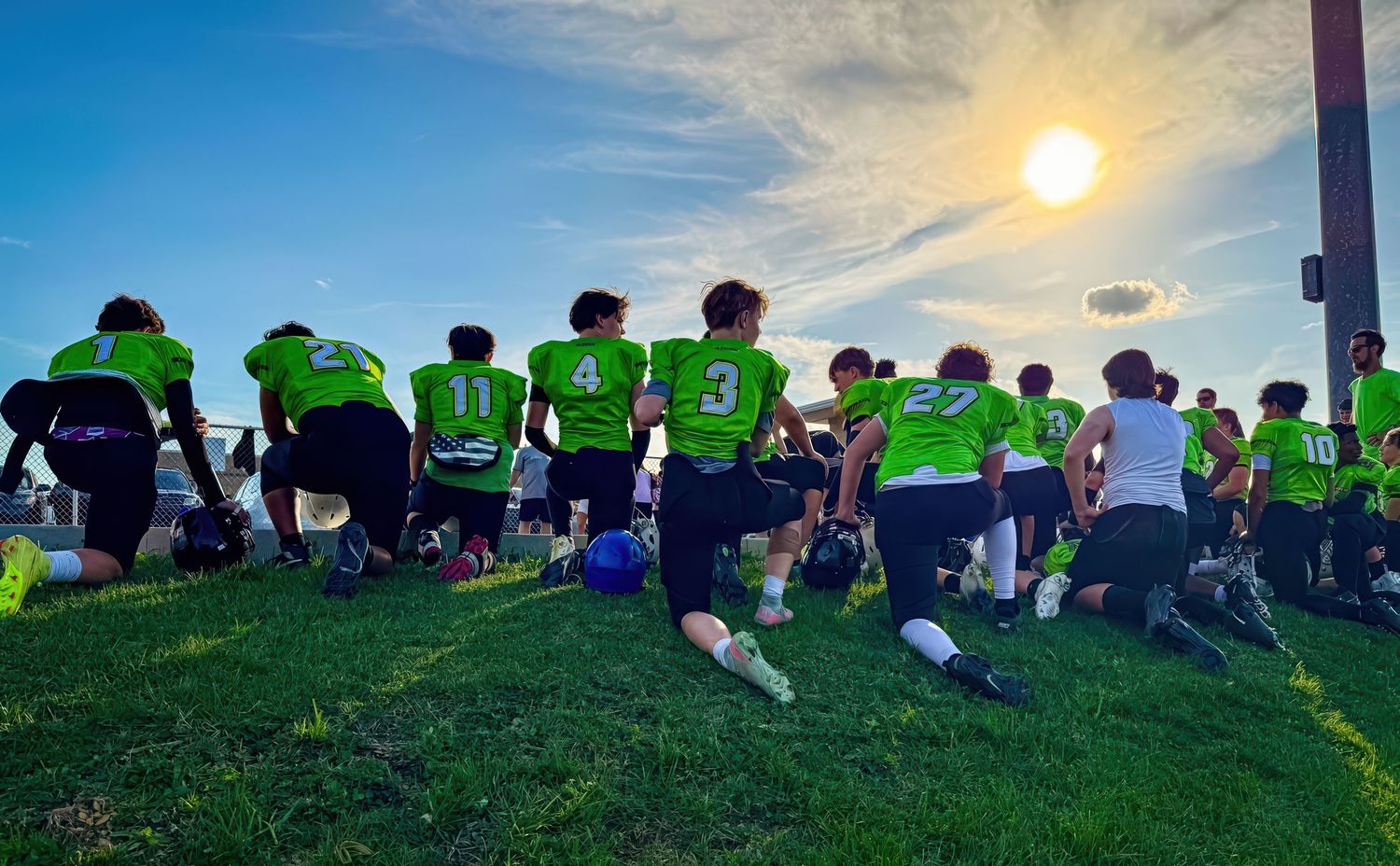 Youth football team in neon green jerseys kneeling together on a grassy hill at sunset.