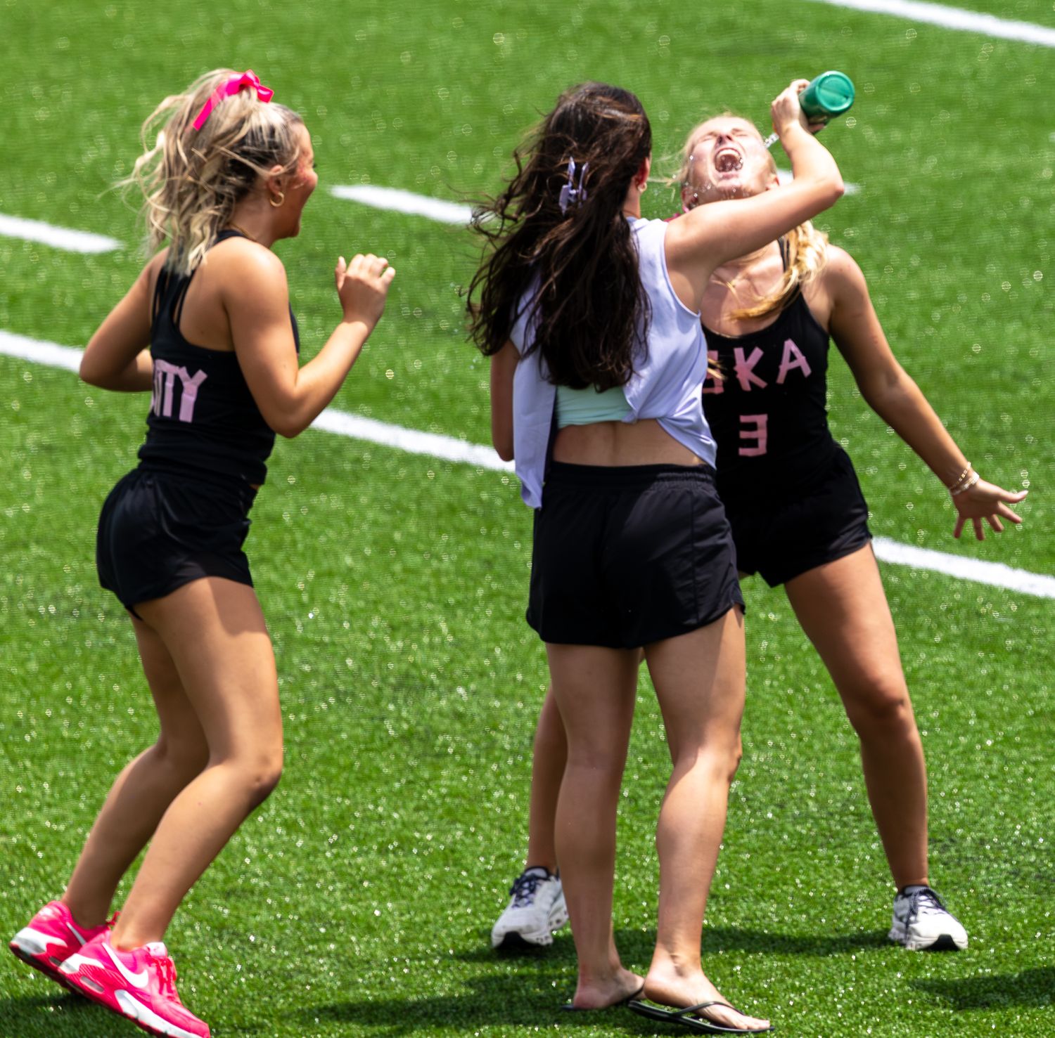 Three young female athletes in pink uniforms celebrate on a green field, one drinking from a water bottle.
