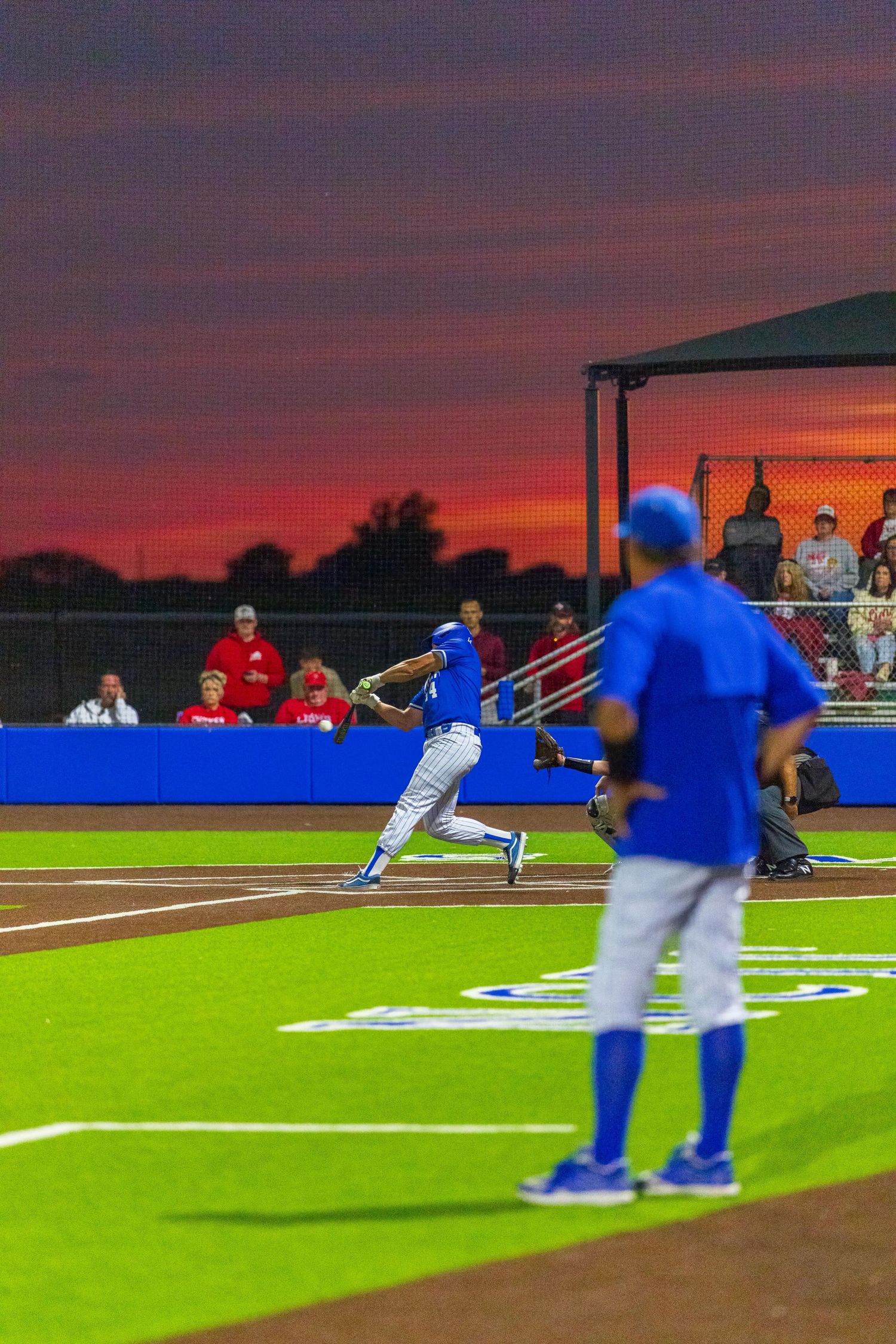 Baseball players in blue uniforms compete under a dramatic red sunset sky at an evening game.