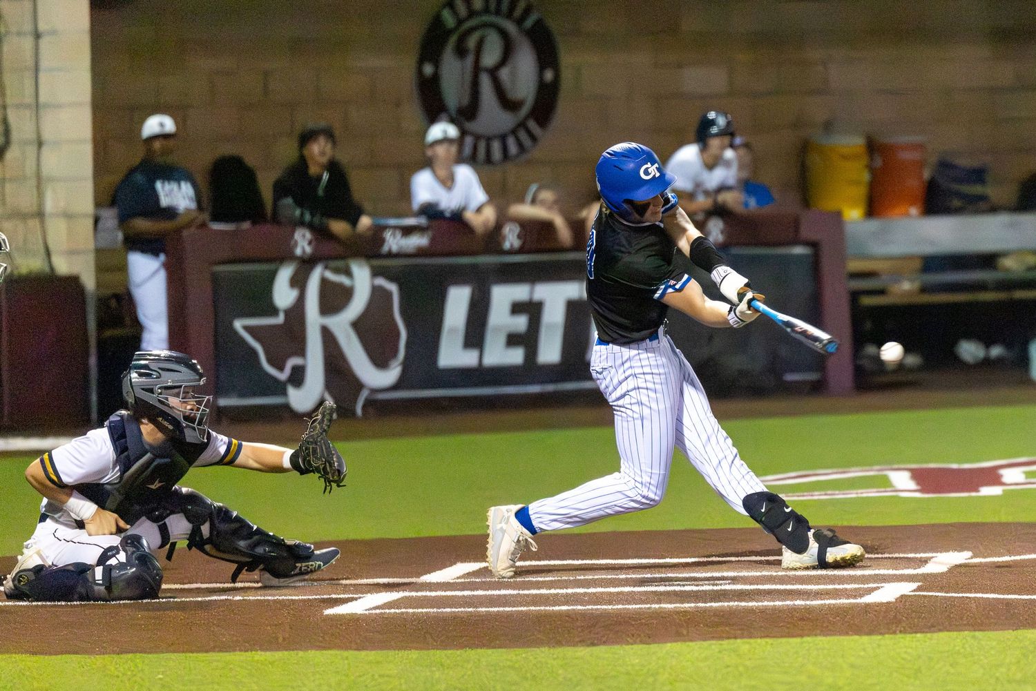 Baseball batter swings at pitch during night game as catcher crouches behind home plate on indoor diamond.