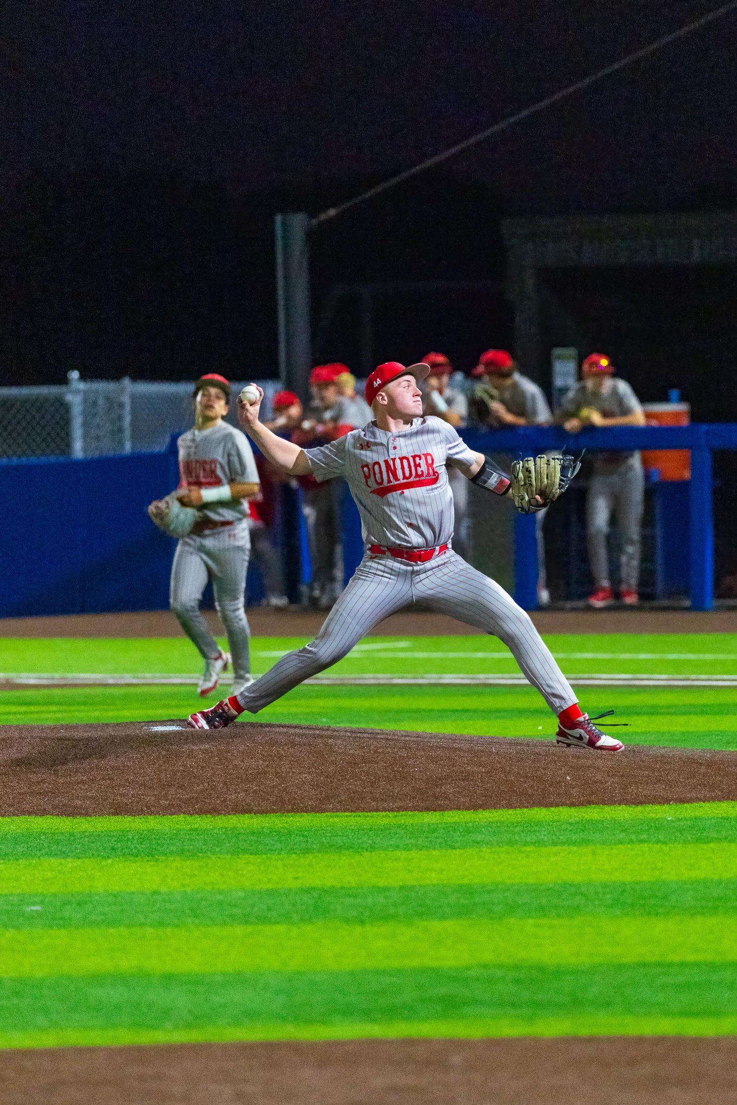 Ponder baseball pitcher in mid-throw during a night game, teammates visible in dugout background.