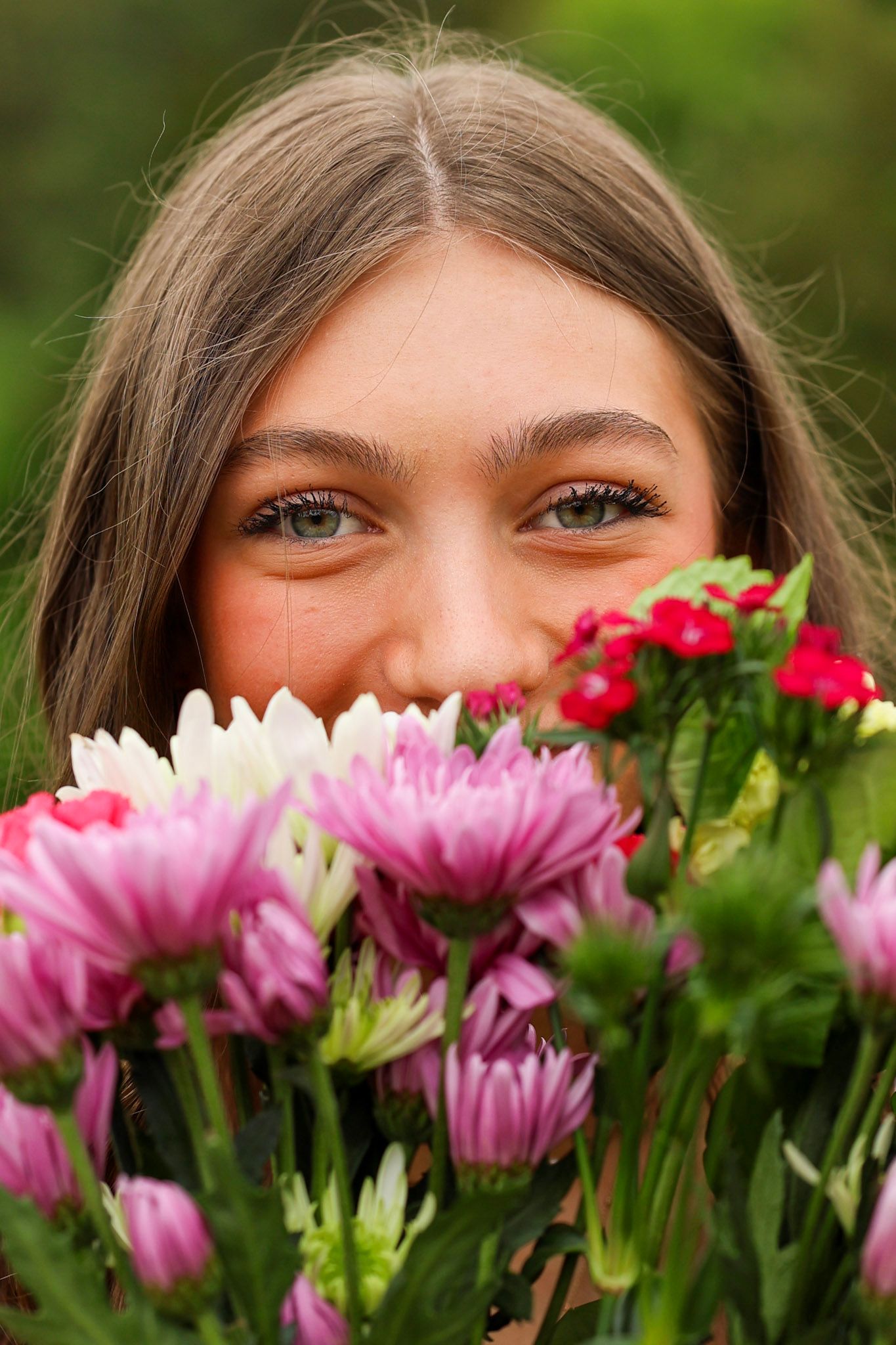 A young woman peeks over a bouquet of colorful pink and red flowers outdoors.