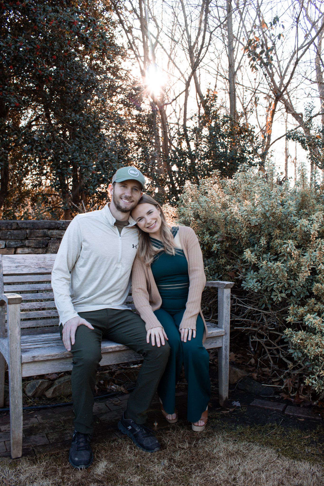 A smiling couple sits together on a wooden bench surrounded by sunlit trees and shrubs.
