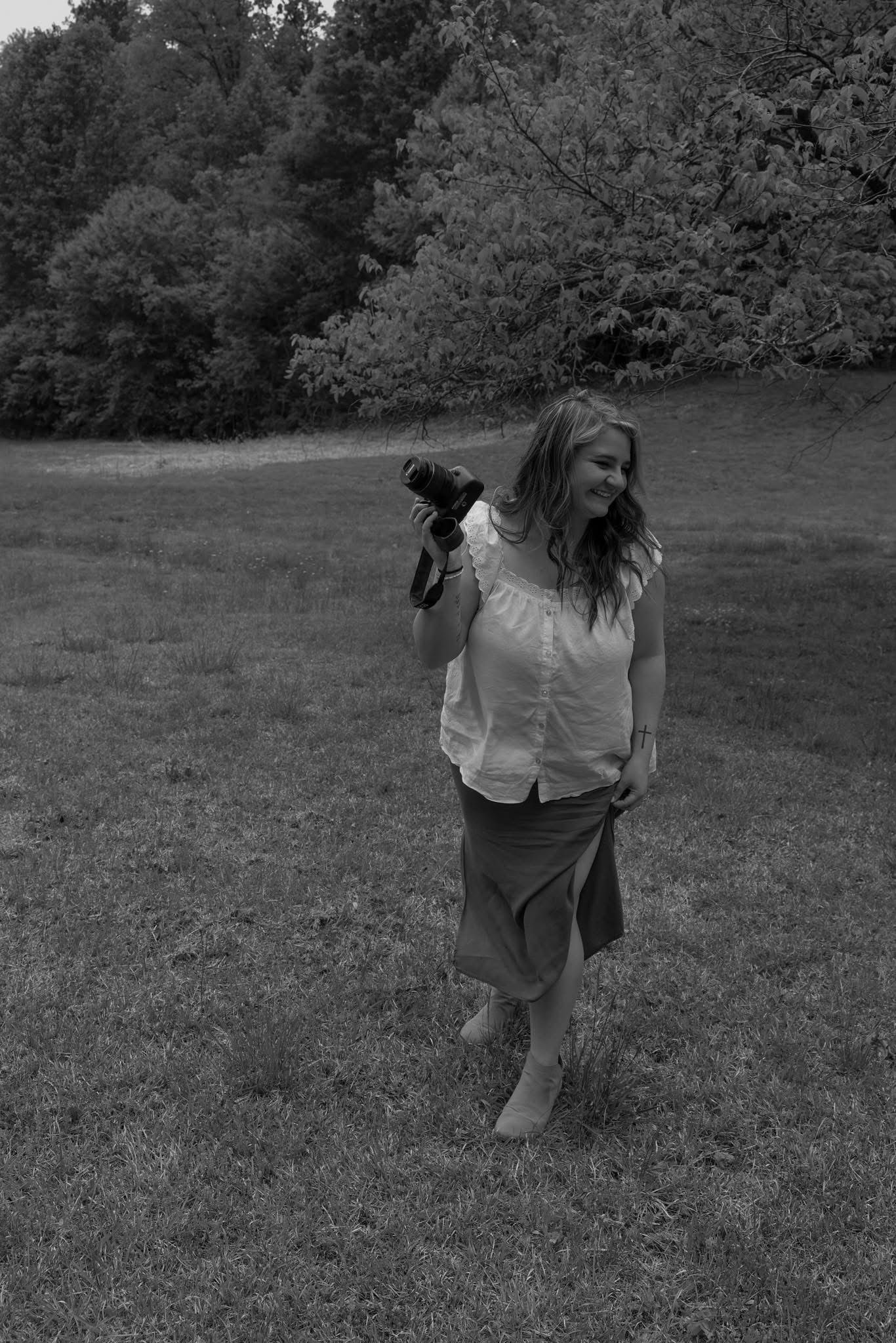 Black and white portrait of a smiling woman holding a camera while walking through a lush green meadow.