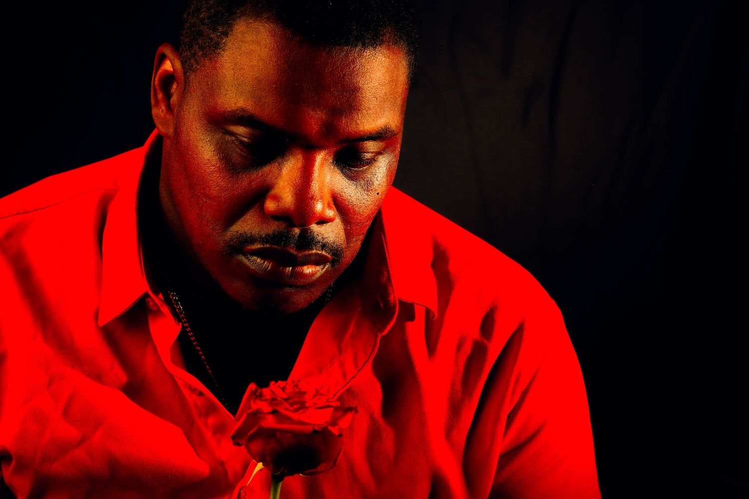 A person in a bright red shirt looks down toward a red rose held close to their face, lit dramatically against a dark background.