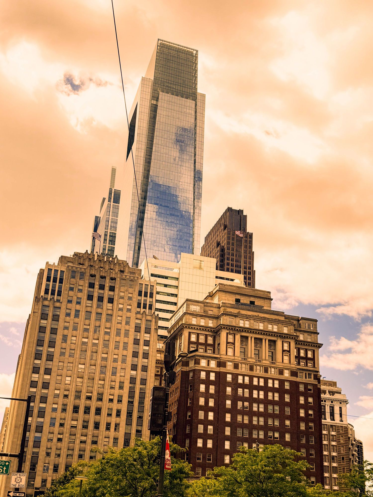 A cityscape featuring a tall glass skyscraper reflecting the sky, surrounded by older brick and stone buildings. Warm golden light fills the