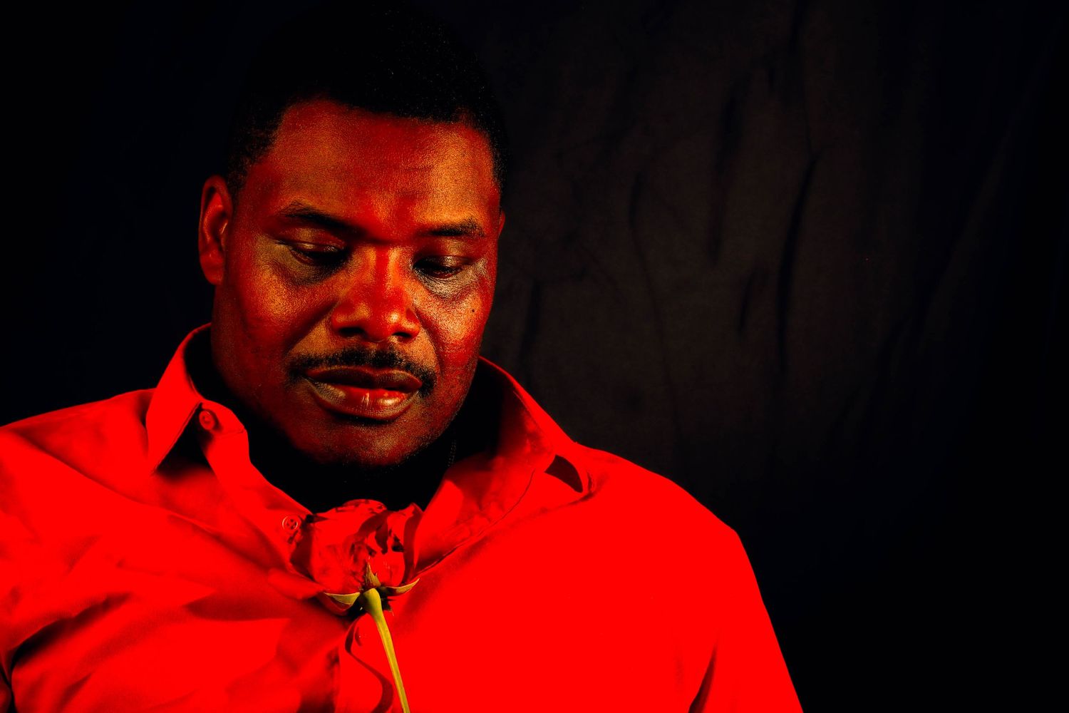 A person wearing a bright red shirt looks downward while a red rose sits near the collar, illuminated against a dark background.