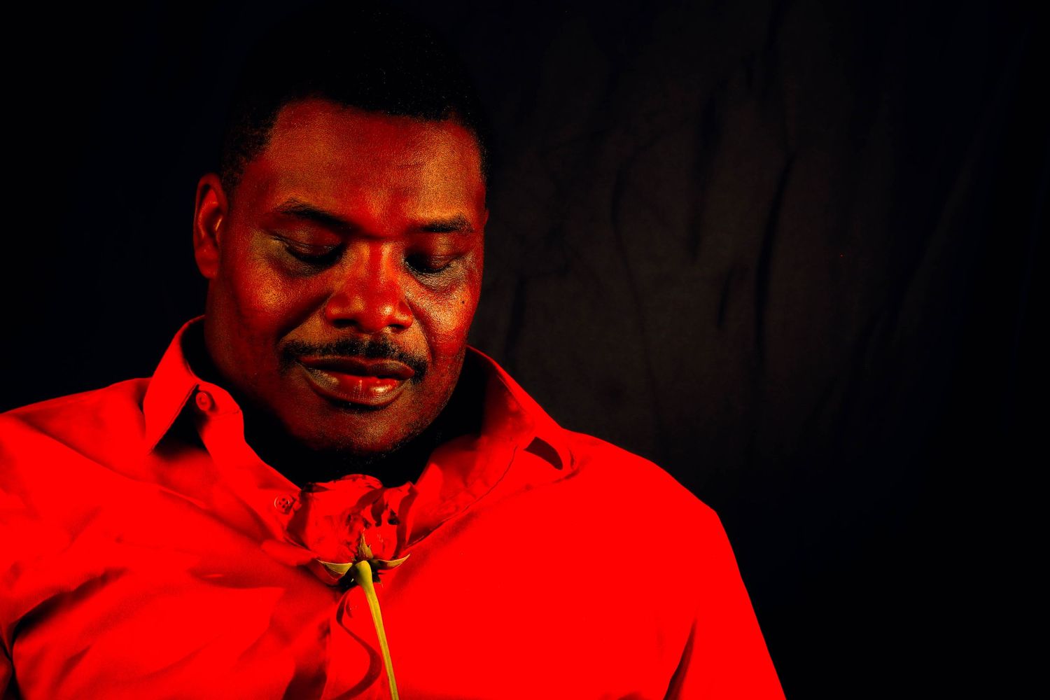 A person in a bright red shirt looks downward with eyes closed as a red rose rests near the collar against a dark background.