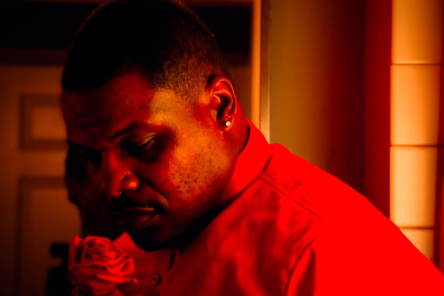 A person in a red shirt holds a red rose close to their face while standing indoors under strong red lighting.