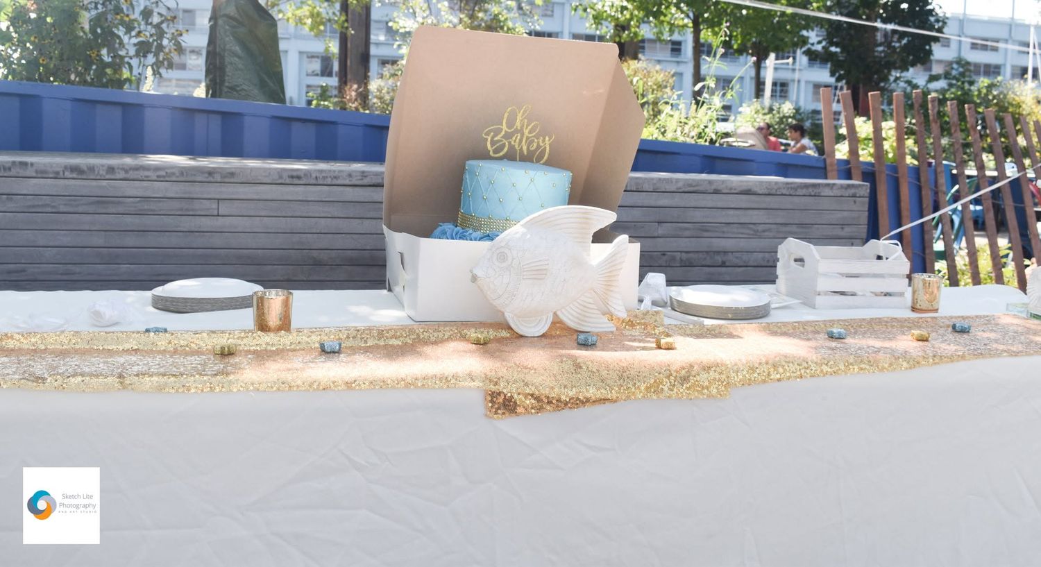 Blue “Oh Baby” cake in an open box on a decorated outdoor table with gold accents, candles, and party details at a baby shower.