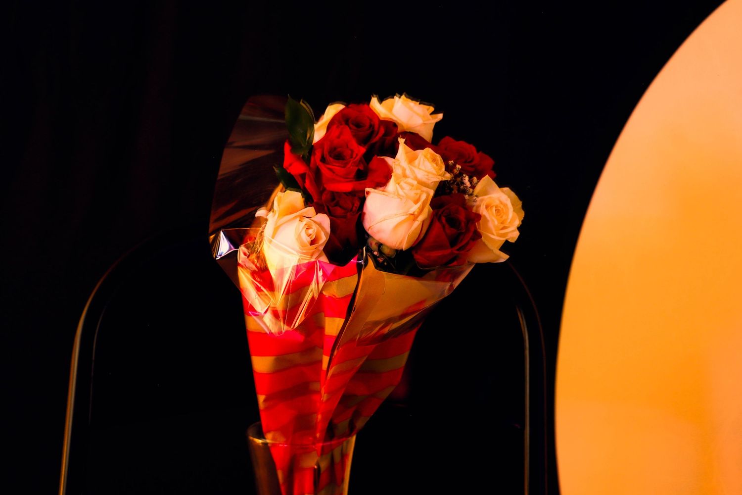 A bouquet of red and white roses wrapped in red‑gold paper, glowing under warm light in a glass vase against a dark, dramatic background.