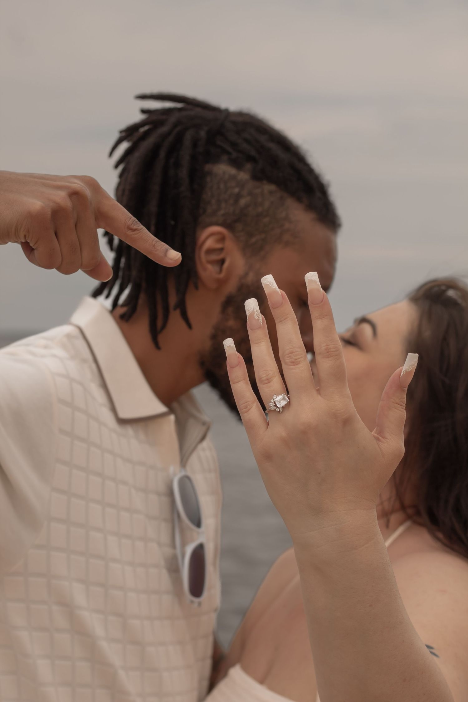 picture of couple at beach, kissing each other, focal point of guy pointing to the girl's hand showing her ring