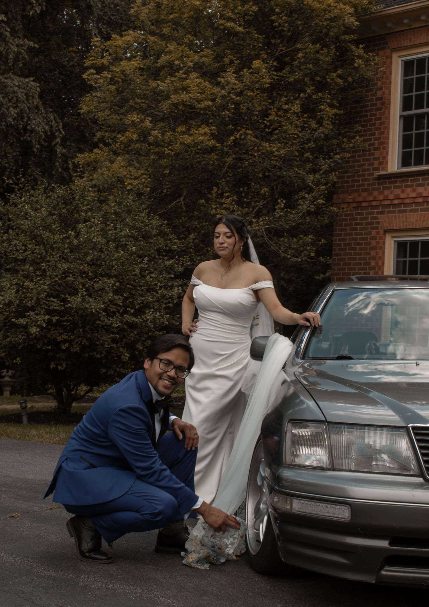 picture of bride and groom sitting and leaning against car. groom is knelt down next to bride