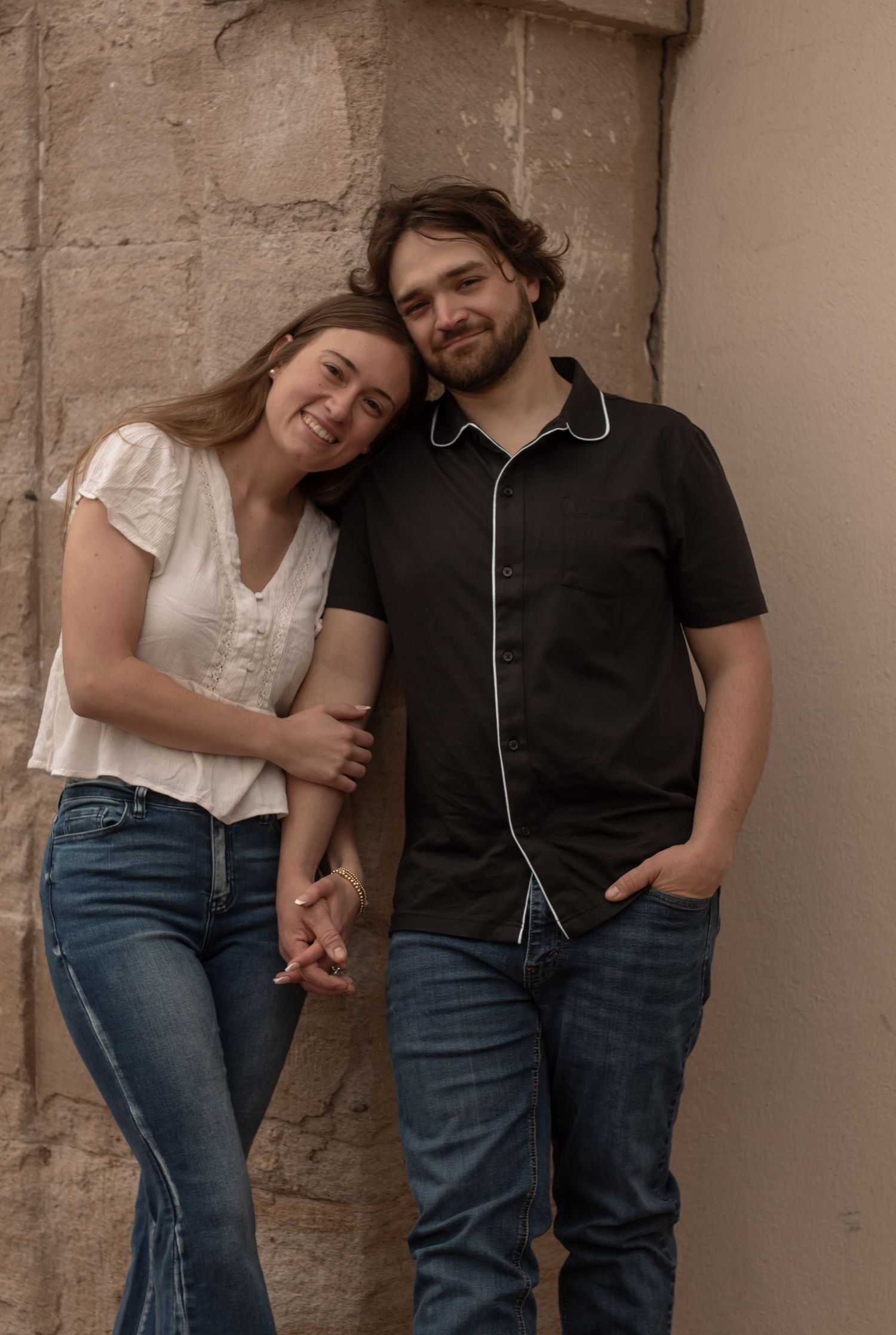 picture of couple, engagement, holding hands and the girl is leaning on the guy's shoulder. they're both leaning up against the brick pillar