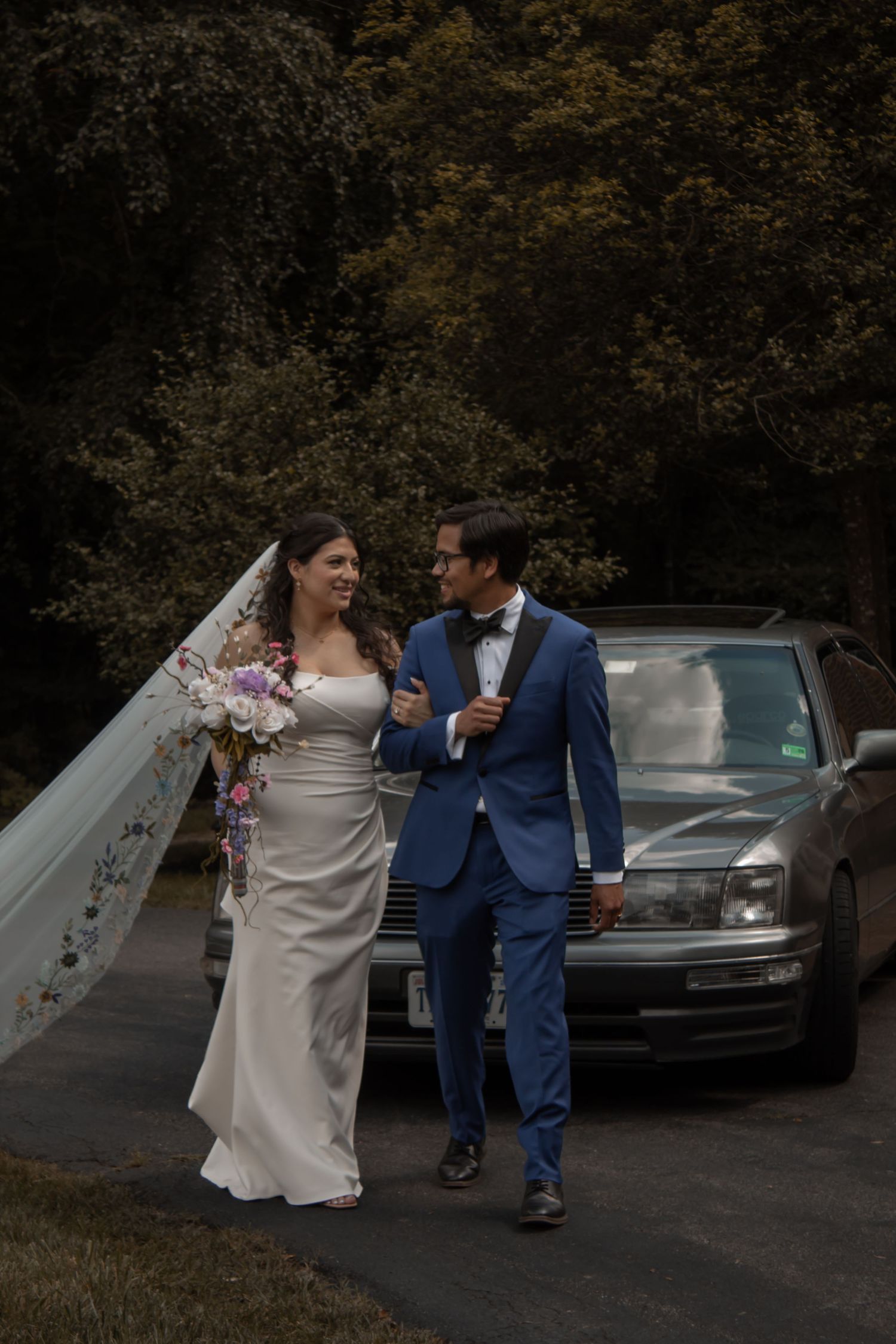 picture of bride and groom looking at each other walking in front of car, bride is holding onto groom's arms