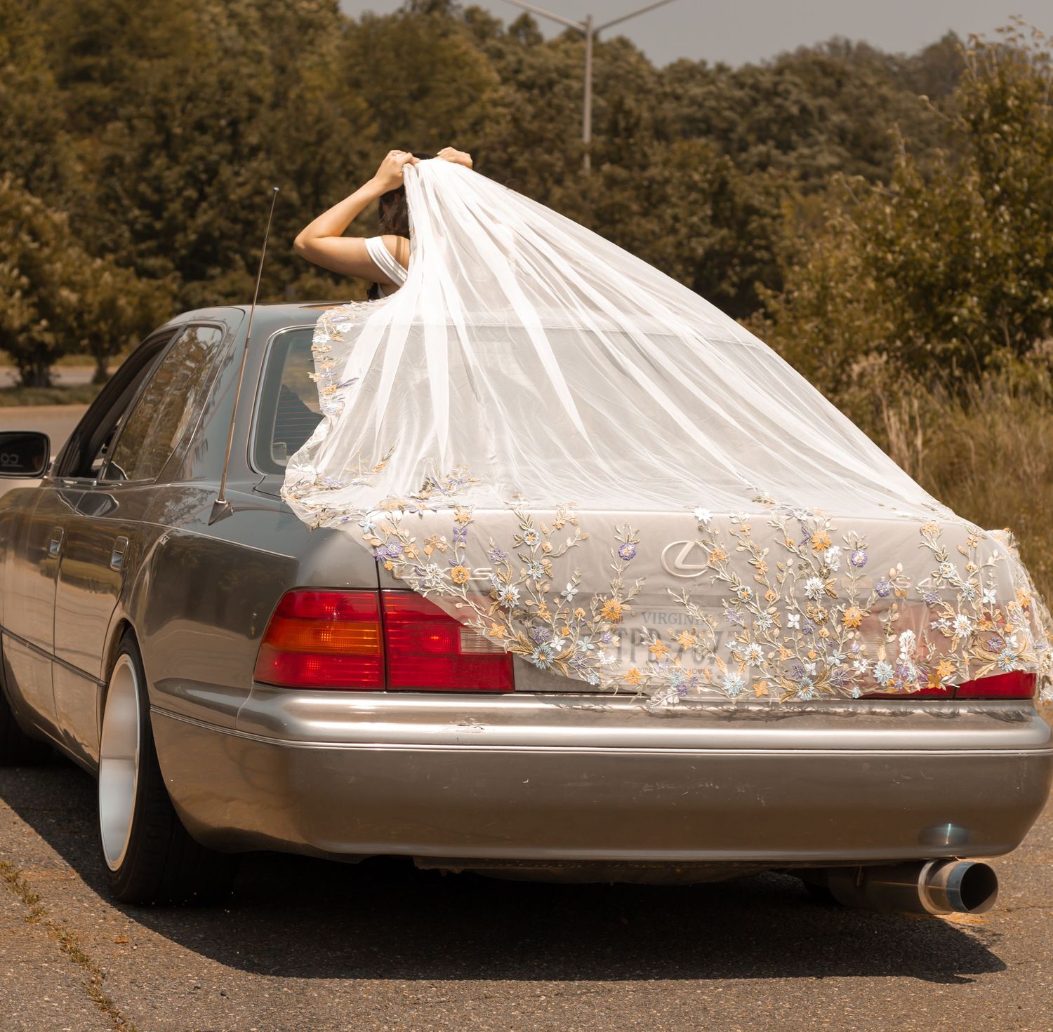 picture of bride sitting in car but poking out through the moon roof, her veil is spread out on top of the car. she's holding her veil.