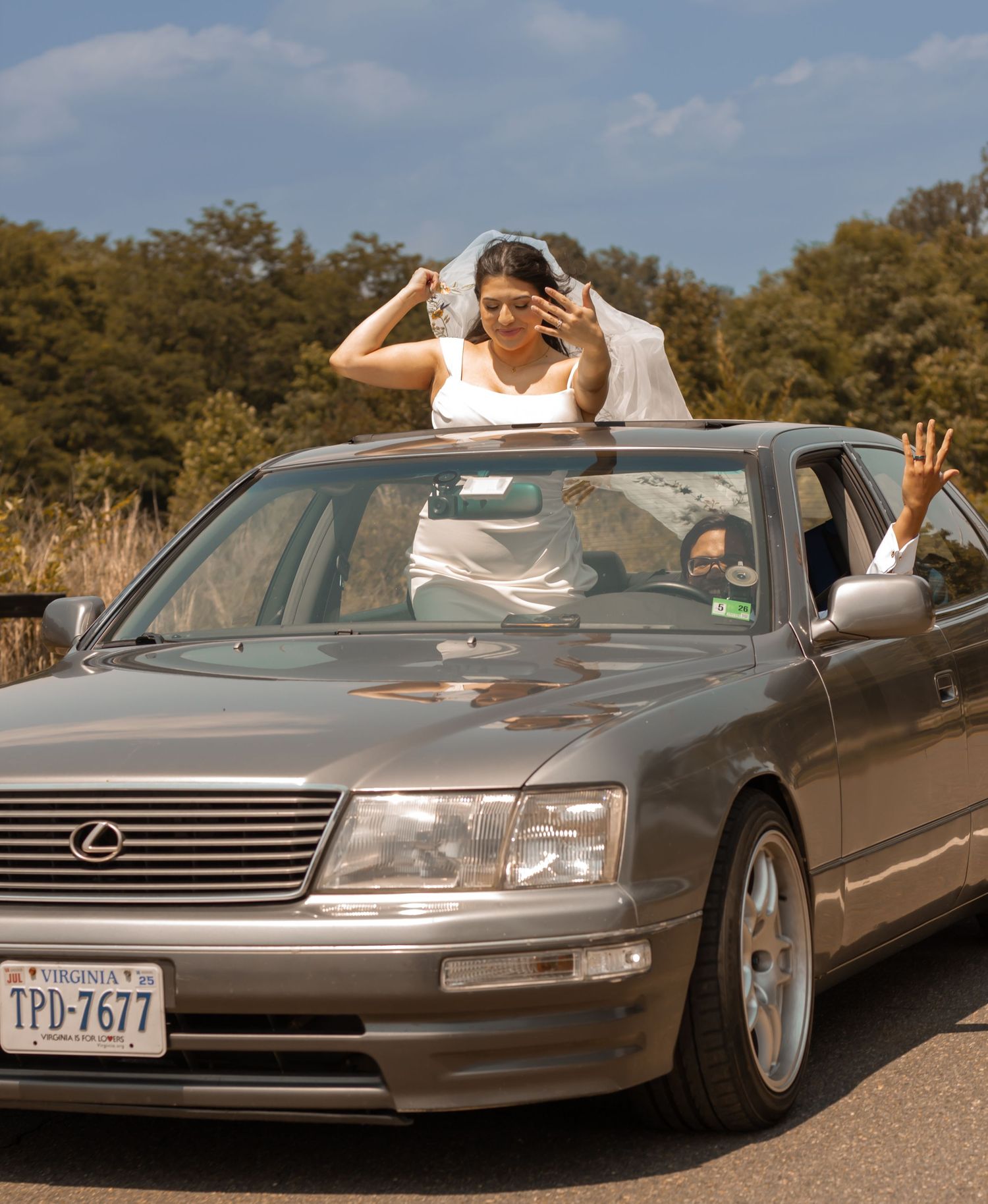 picture of bride and groom in the car. groom is on driver seat, showing his hands, bride poking up the moon roof and showing her hand too