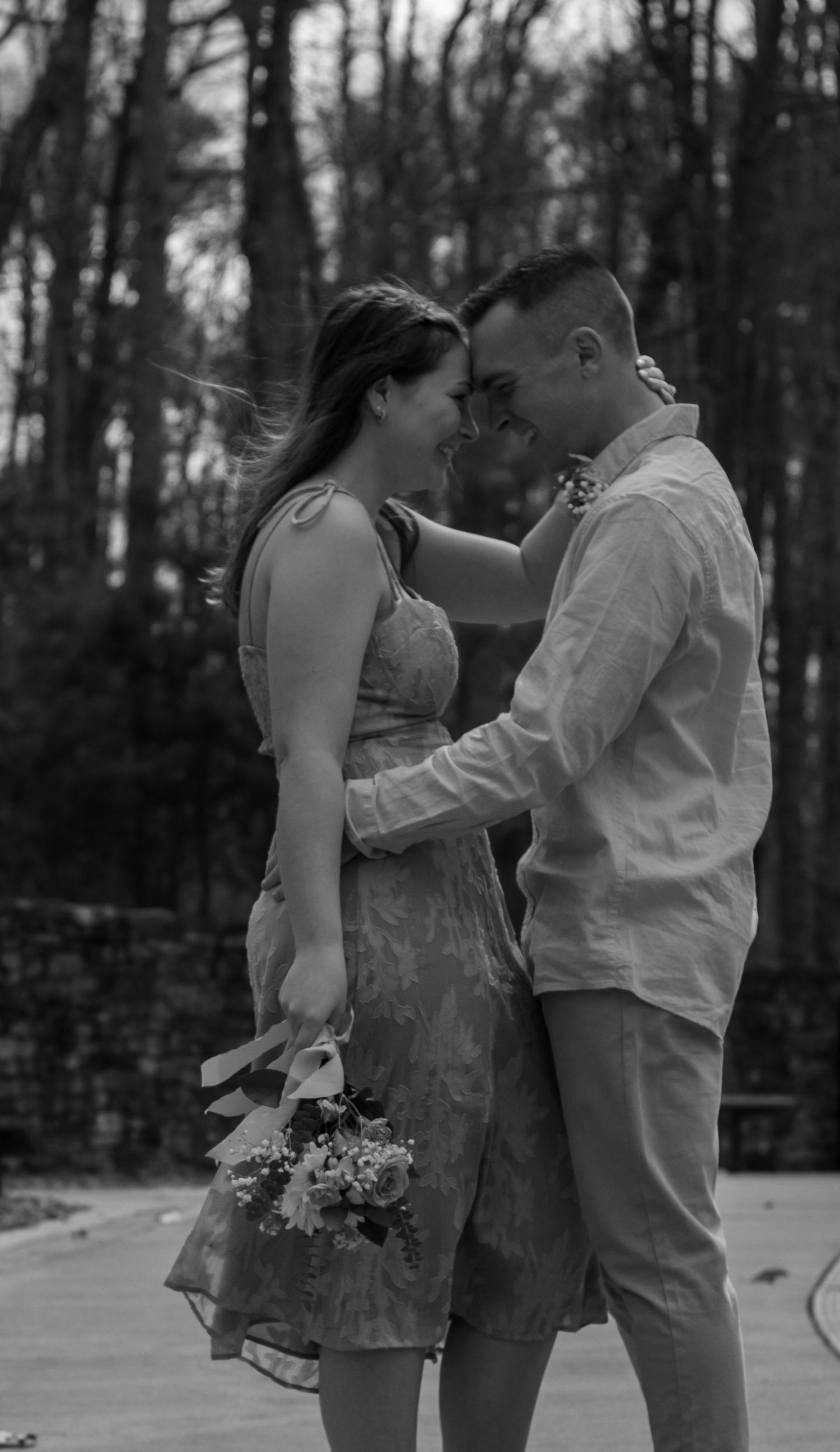 picture of couple in woods, holding each other and looking at each other. girl is holding a bouquet of flowers