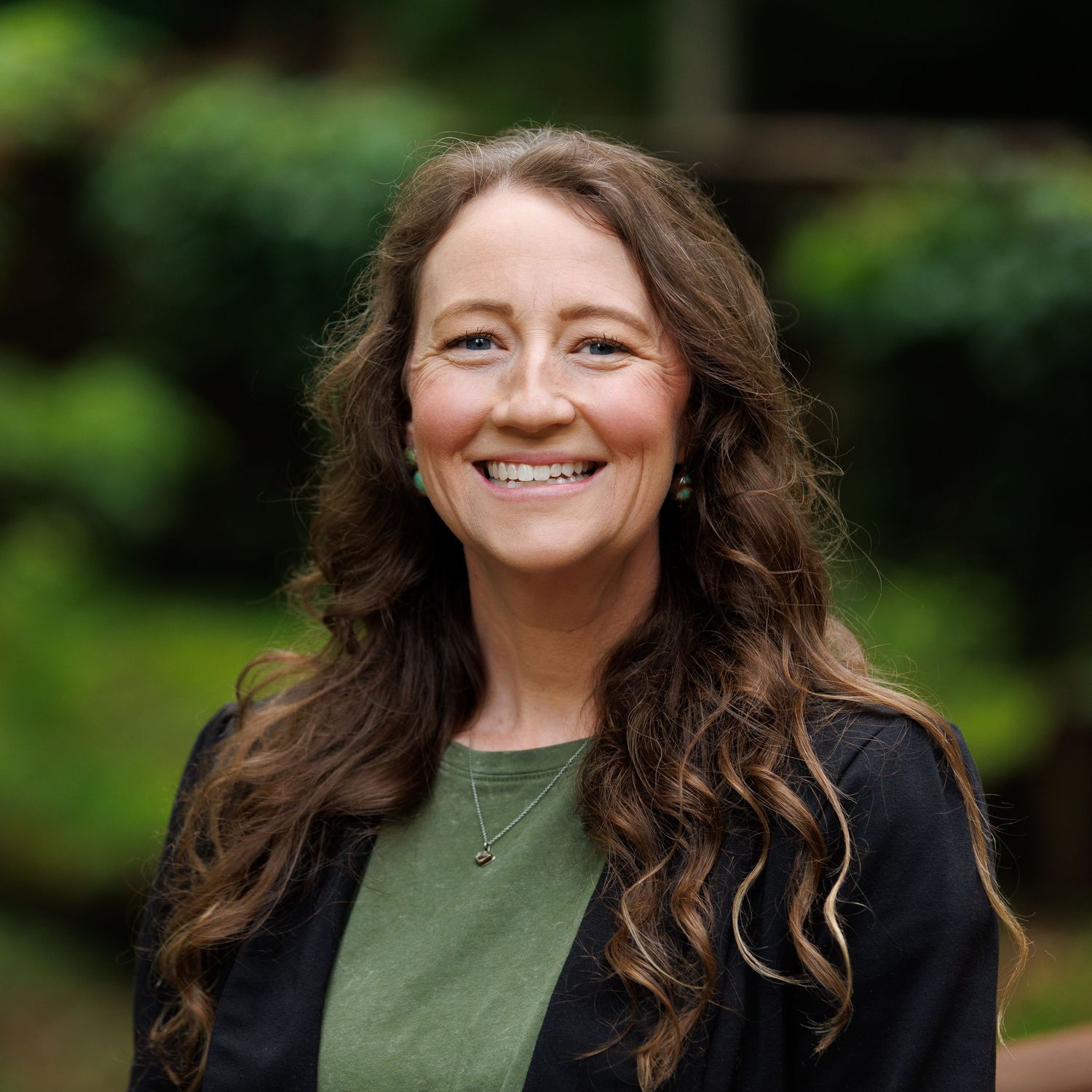 Smiling woman with long wavy hair wearing a green shirt and black blazer outdoors.