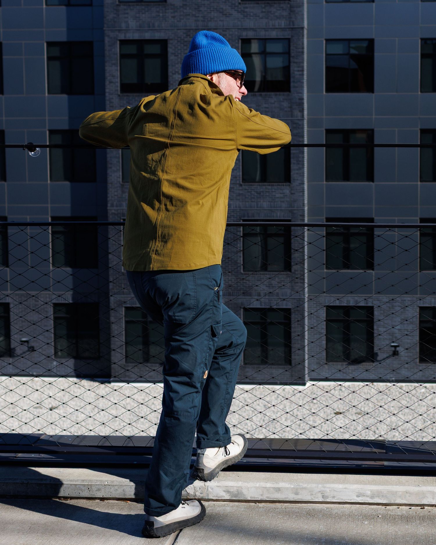Rear view of man in olive jacket and blue beanie leaning on urban rooftop railing.