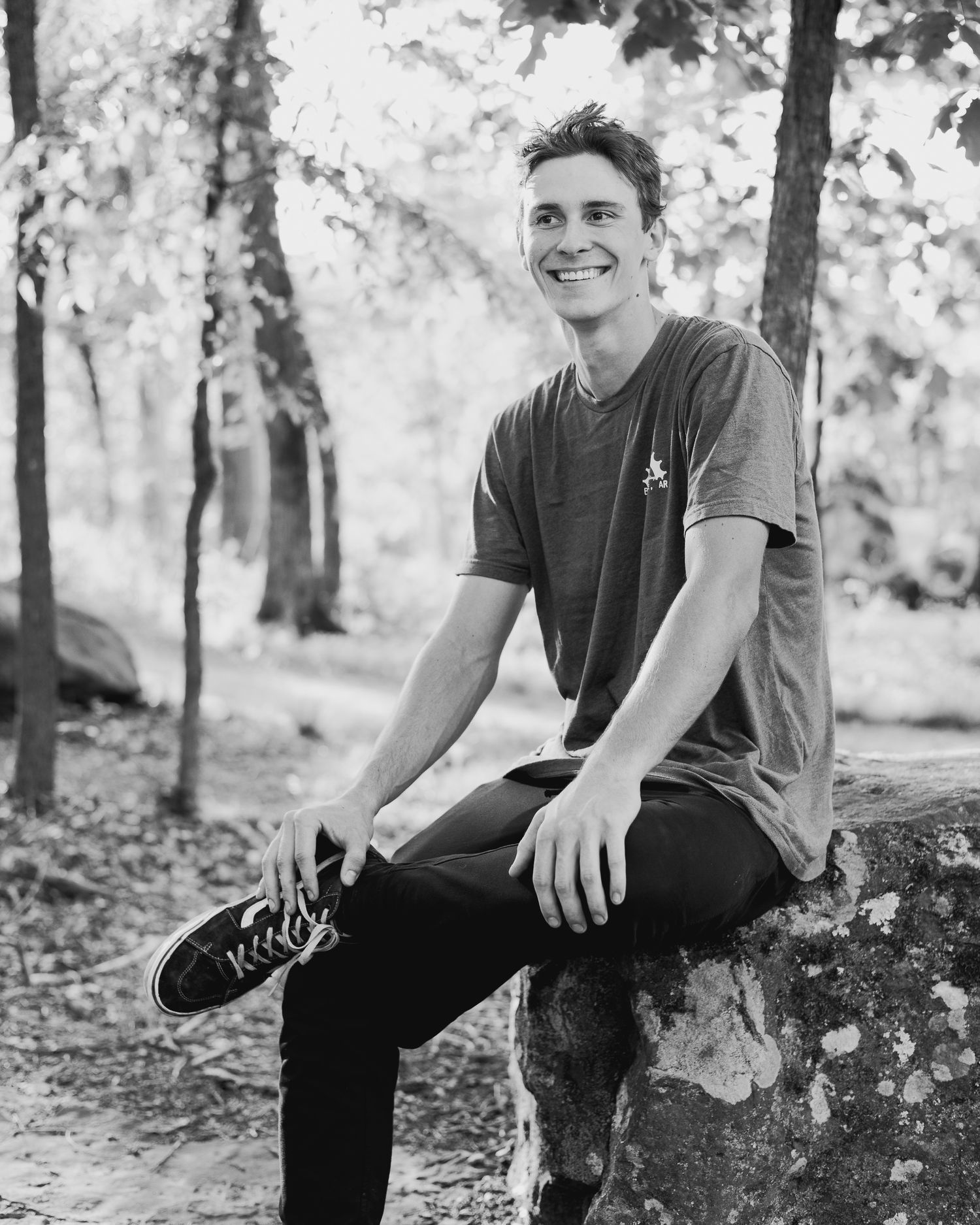 Black and white portrait of a smiling young man sitting on a rock in a sunlit forest.
