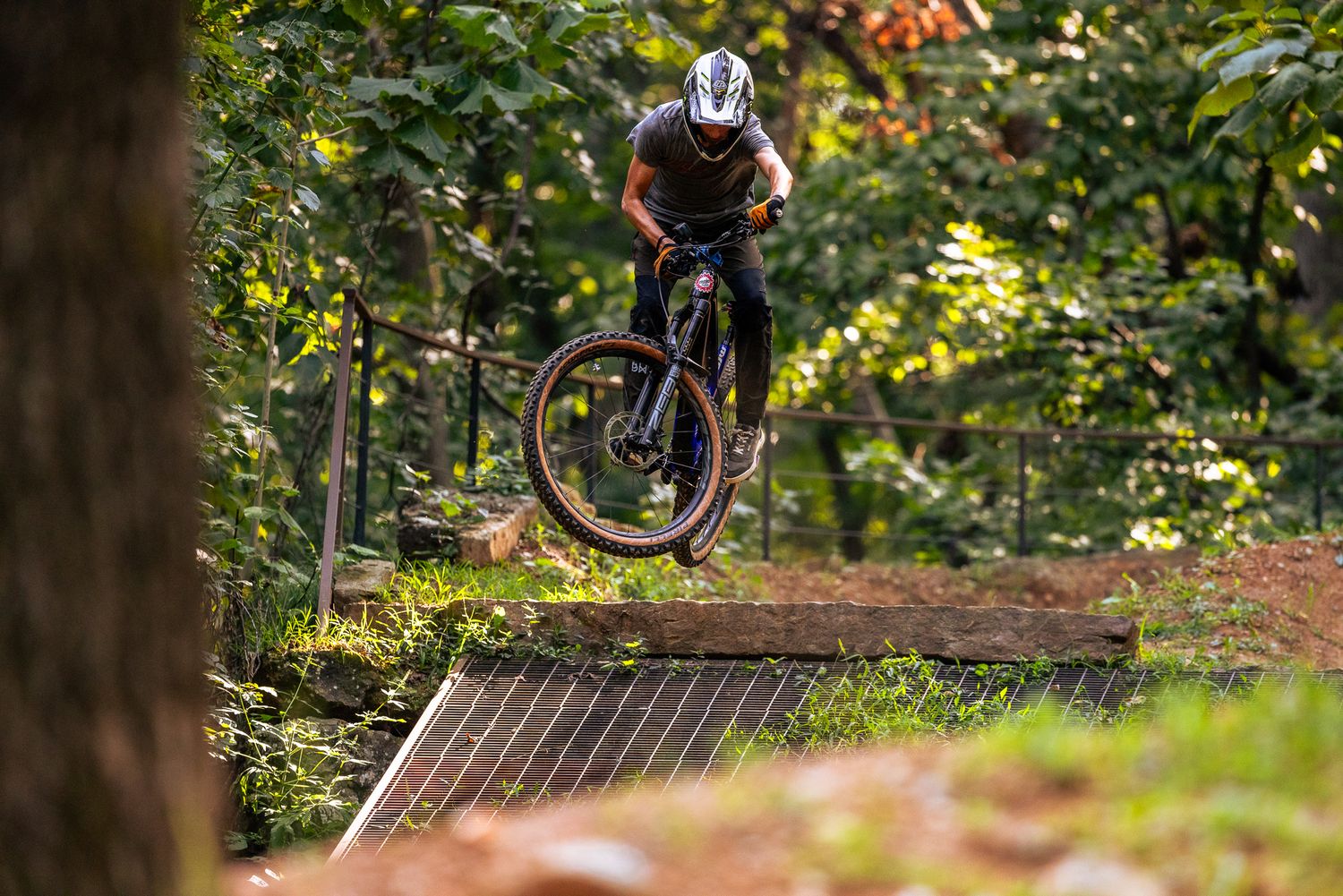 Mountain biker performs aerial jump over wooden ramp feature in forest trail setting.
