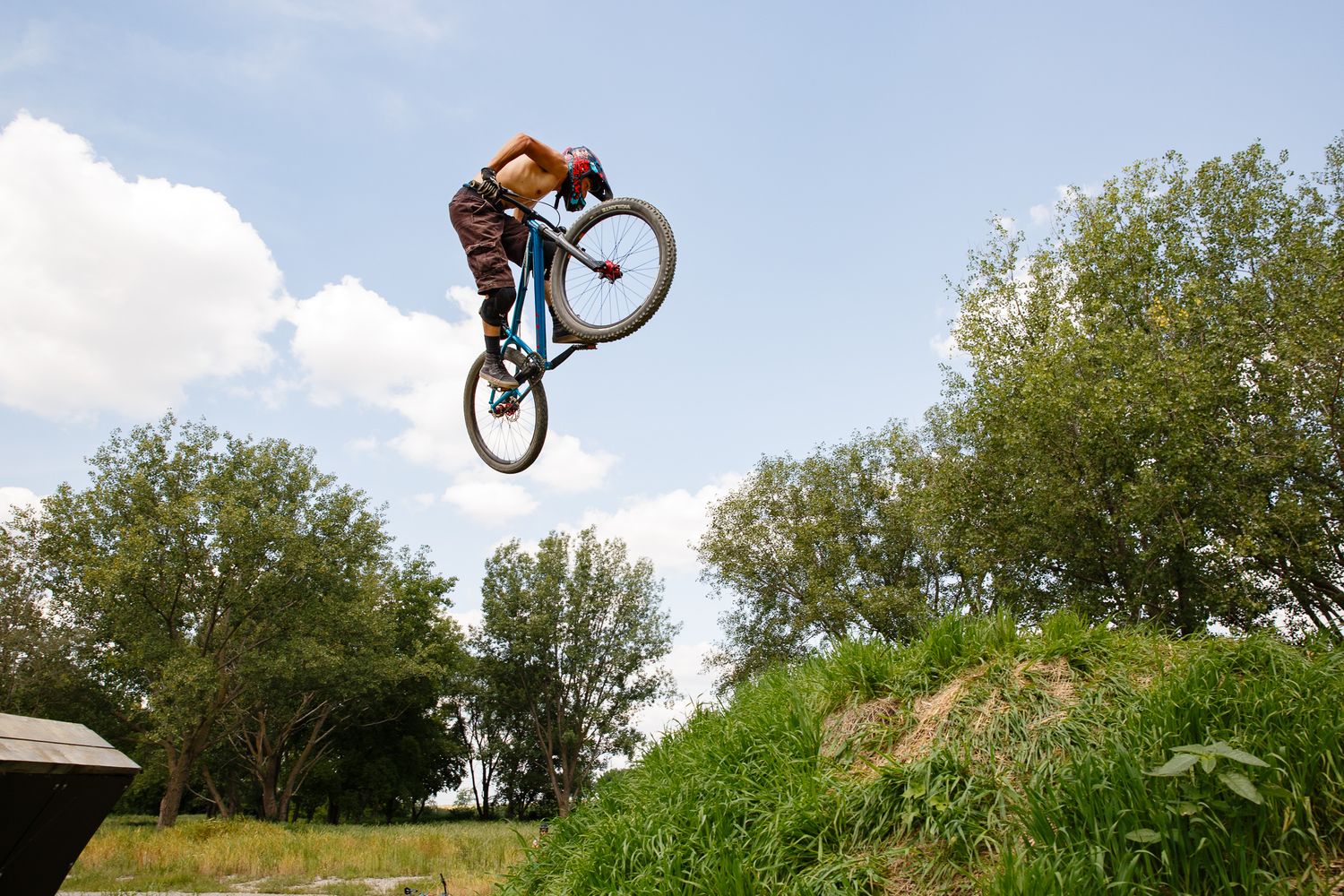 BMX rider performs a high aerial jump trick over a dirt mound against a bright blue summer sky.