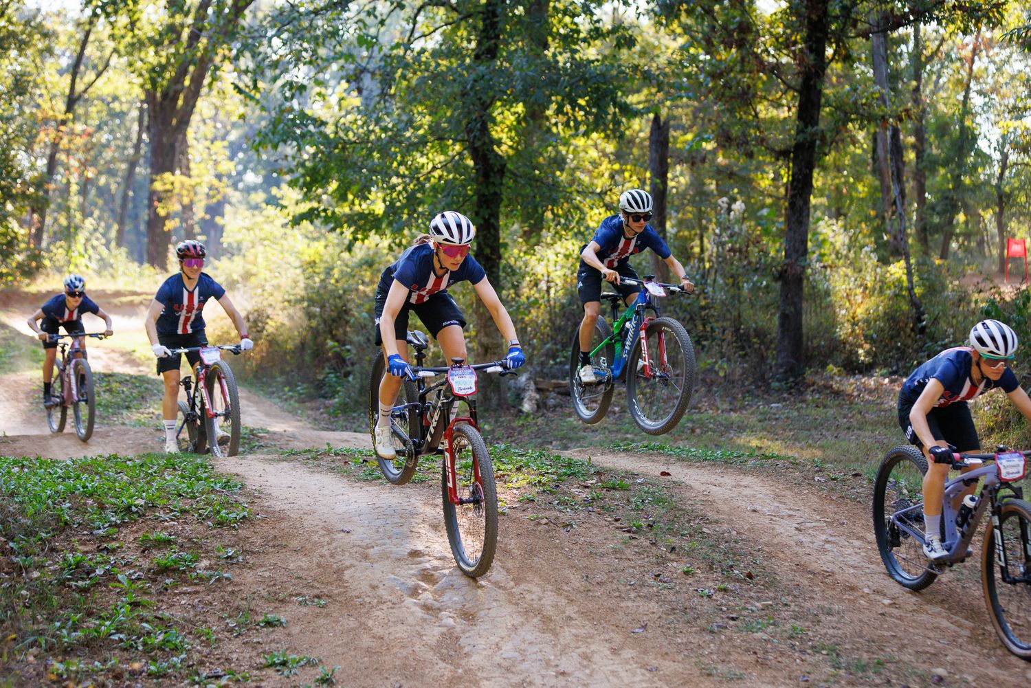Group of mountain bikers racing together on a dusty dirt trail through a sunlit autumn forest.