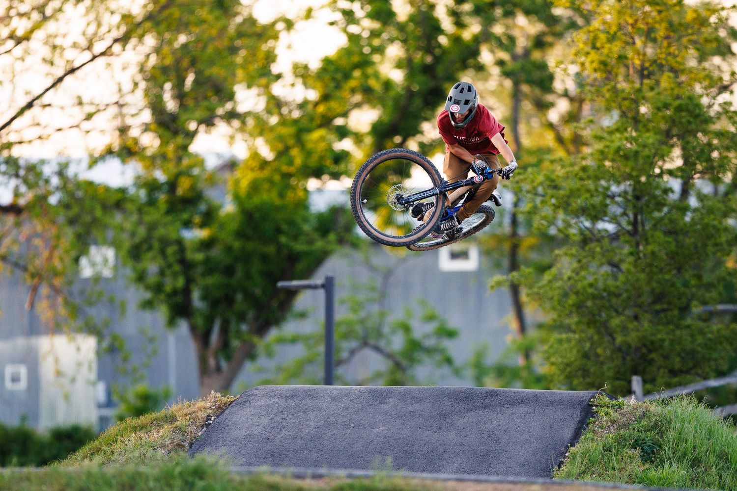 Cyclist performs a big air jump on a mountain bike at an outdoor pump track surrounded by green trees.
