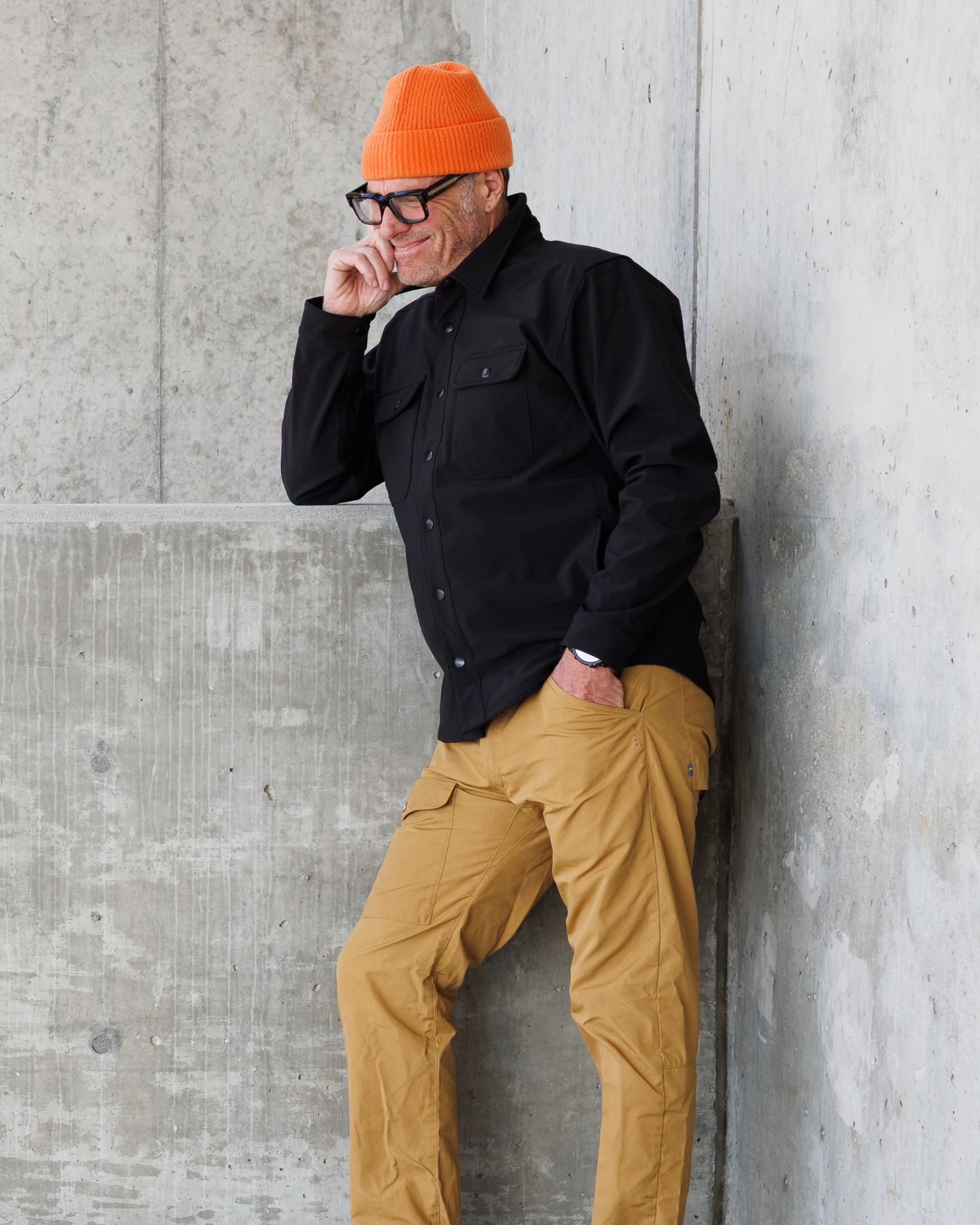 Man in black shirt jacket and orange beanie leaning against concrete wall with khaki cargo pants.