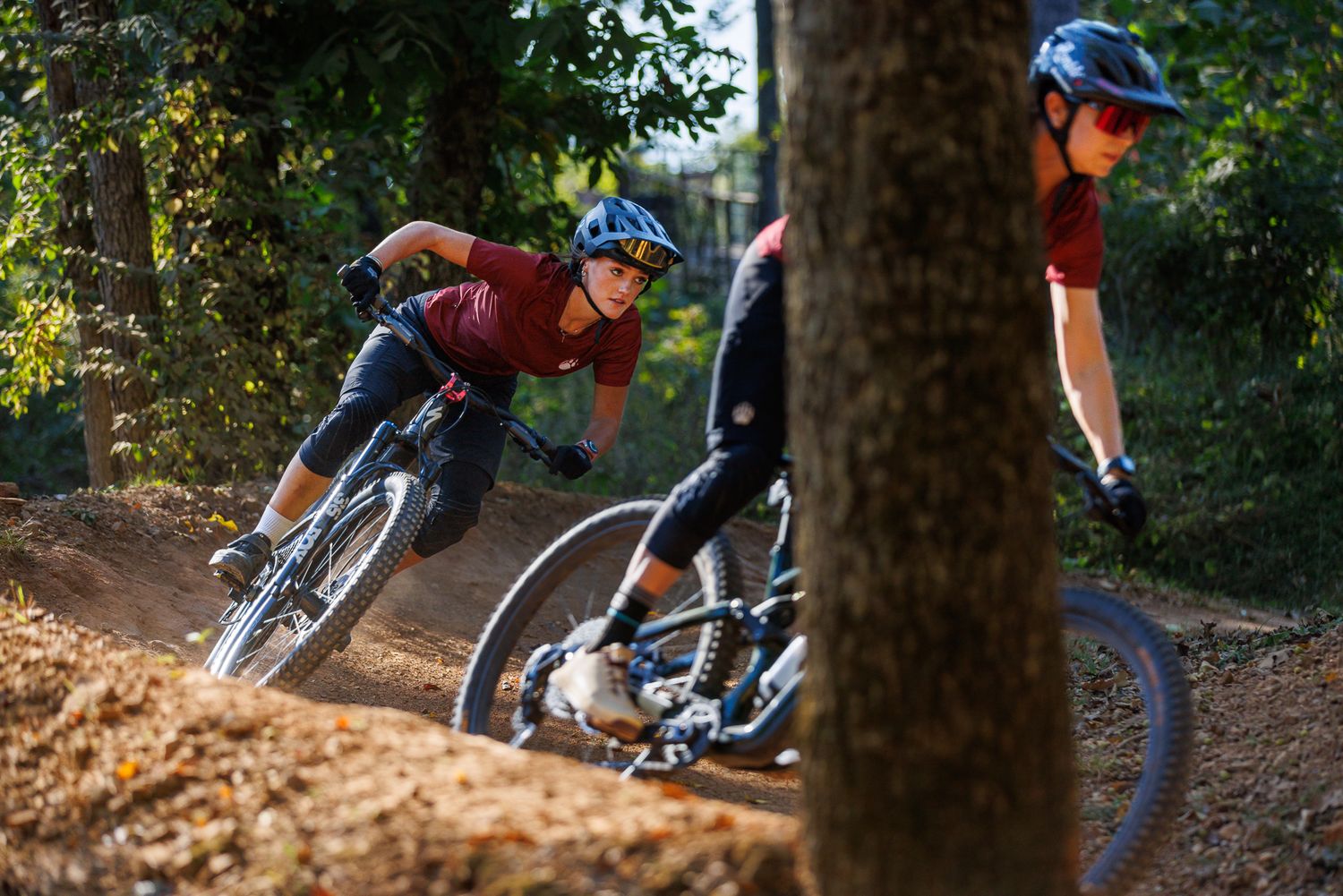 Two mountain bikers leaning into a sharp turn on a dirt trail through a forest.