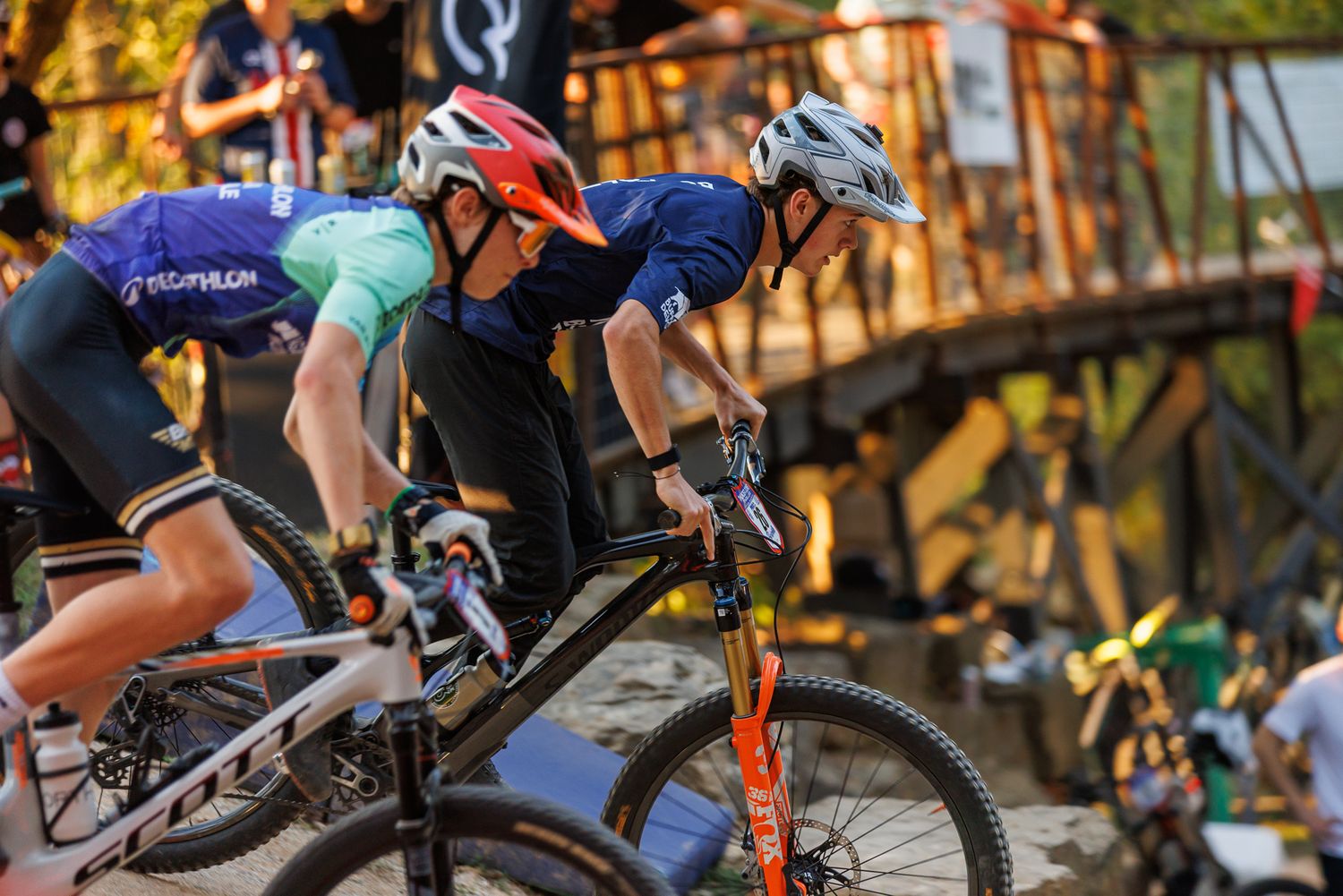 Three mountain bikers compete in a cross-country race near a wooden bridge at a cycling event.