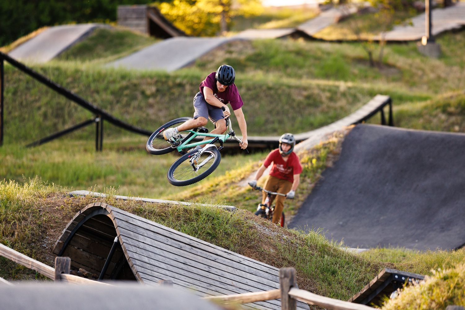 Two cyclists perform tricks at an outdoor bike pump track park on a sunny day.