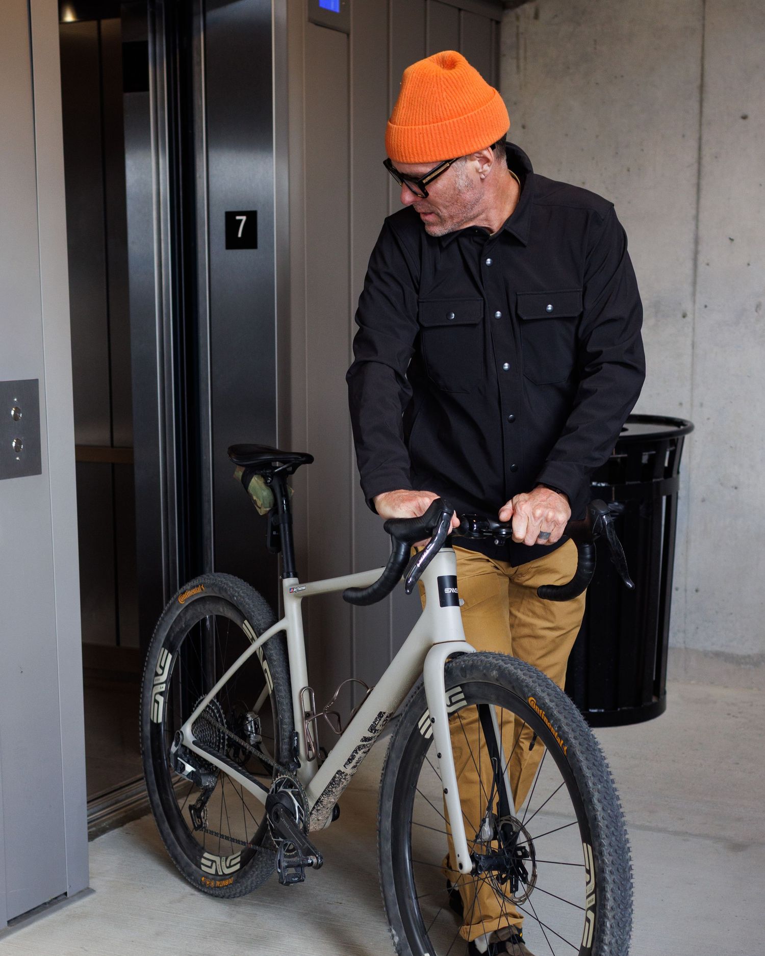 Man in black jacket and orange beanie loading bicycle into elevator with cargo pants.