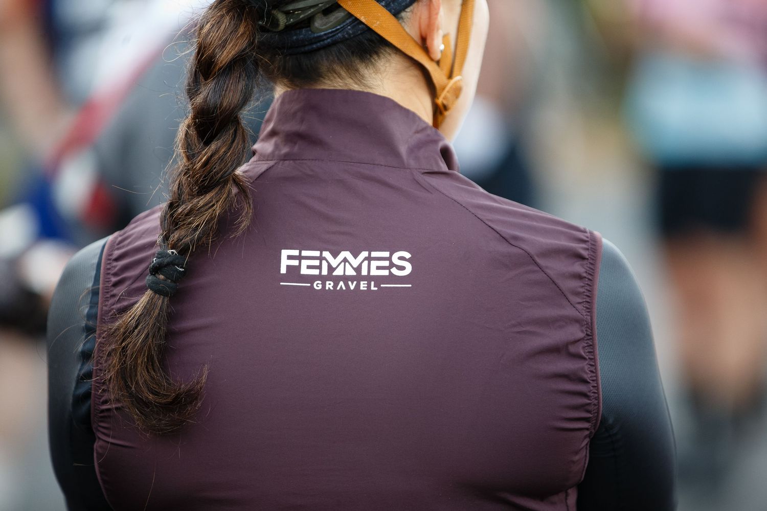 Woman cyclist wearing a burgundy Femmes Gravel vest with logo, hair in braid, helmet straps visible.