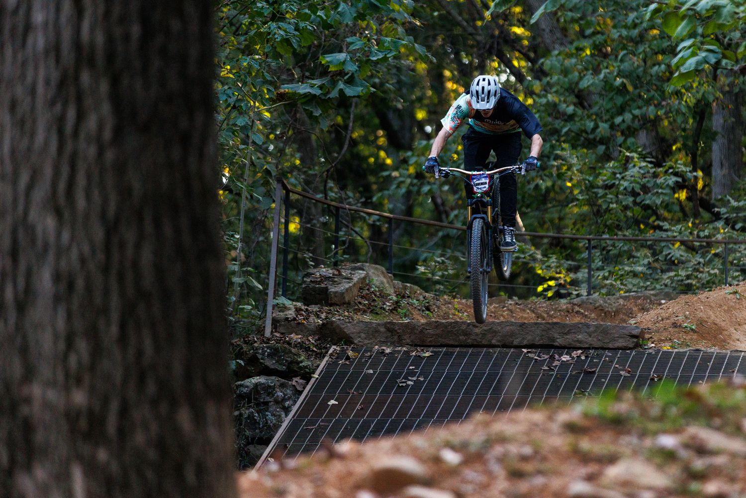 Mountain biker descending a wooden bridge feature on a dark forest trail during a race.