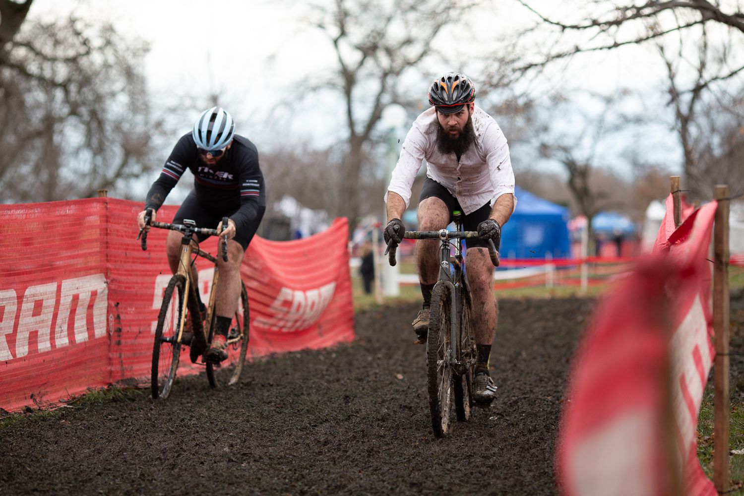 Two cyclists compete in a muddy cyclocross race on a grass course marked with red and white barriers.