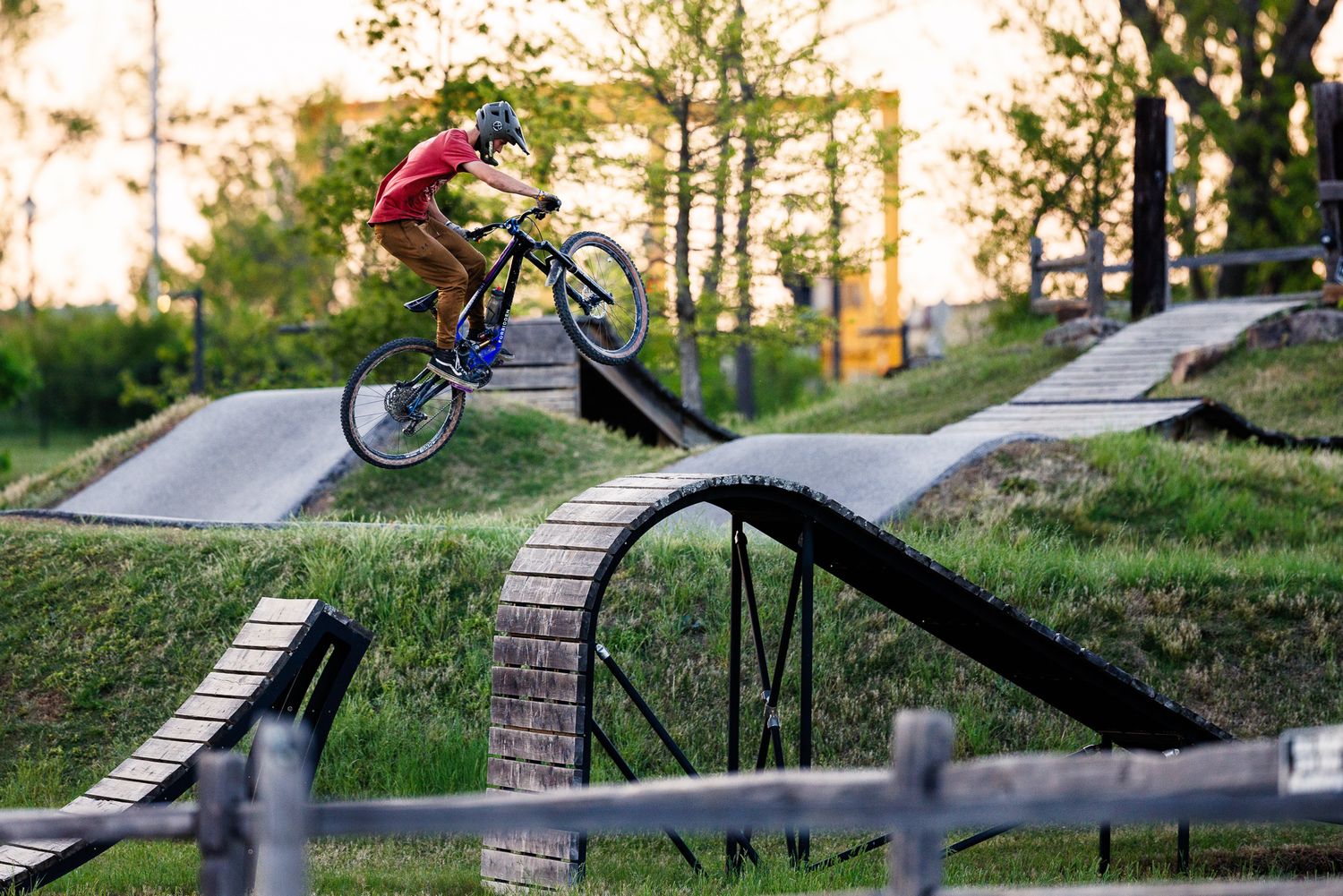 Mountain biker launches off a jump on a wooden pump track course surrounded by lush greenery.