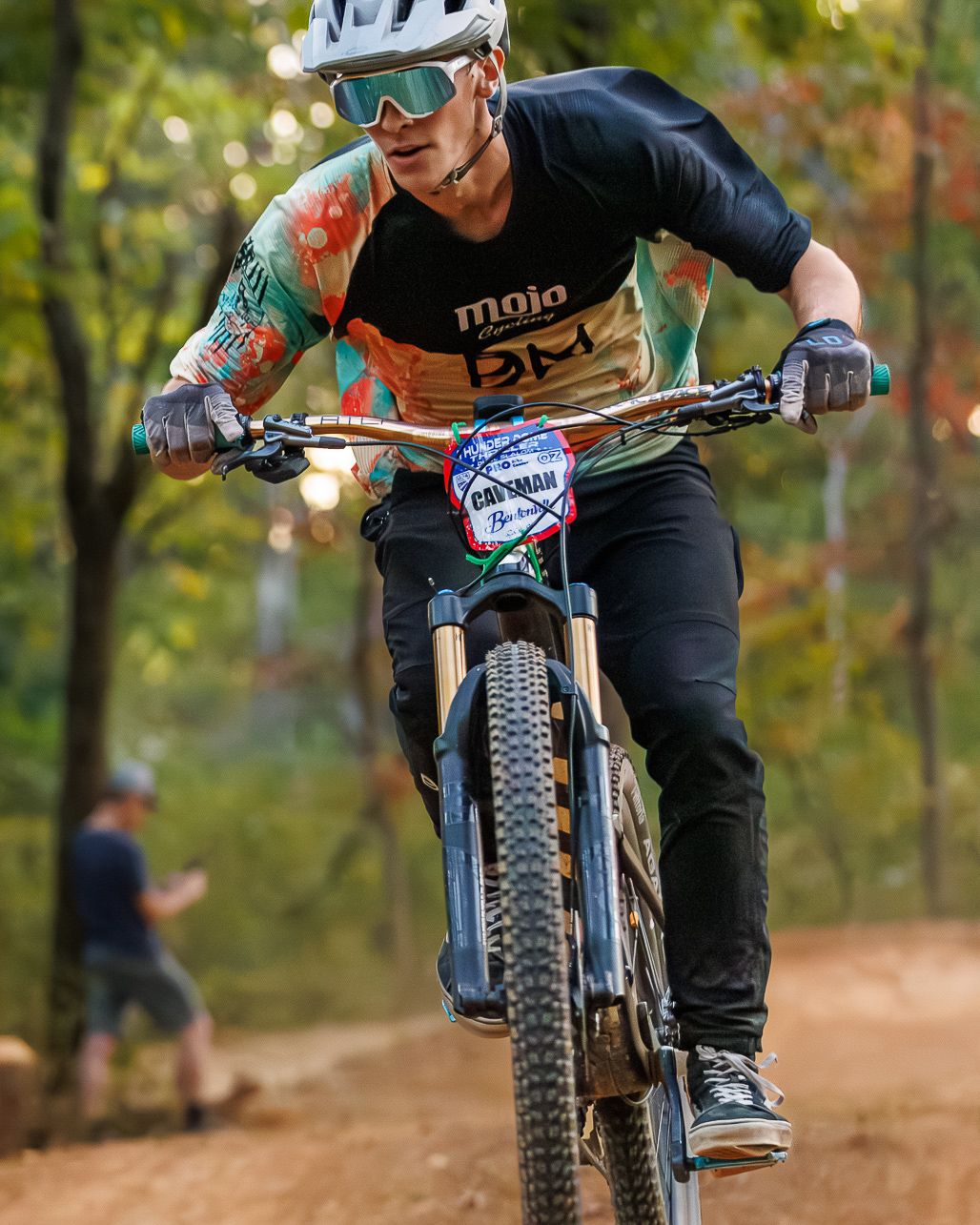 Mountain biker in Mojo jersey races downhill on dirt trail through autumn forest.